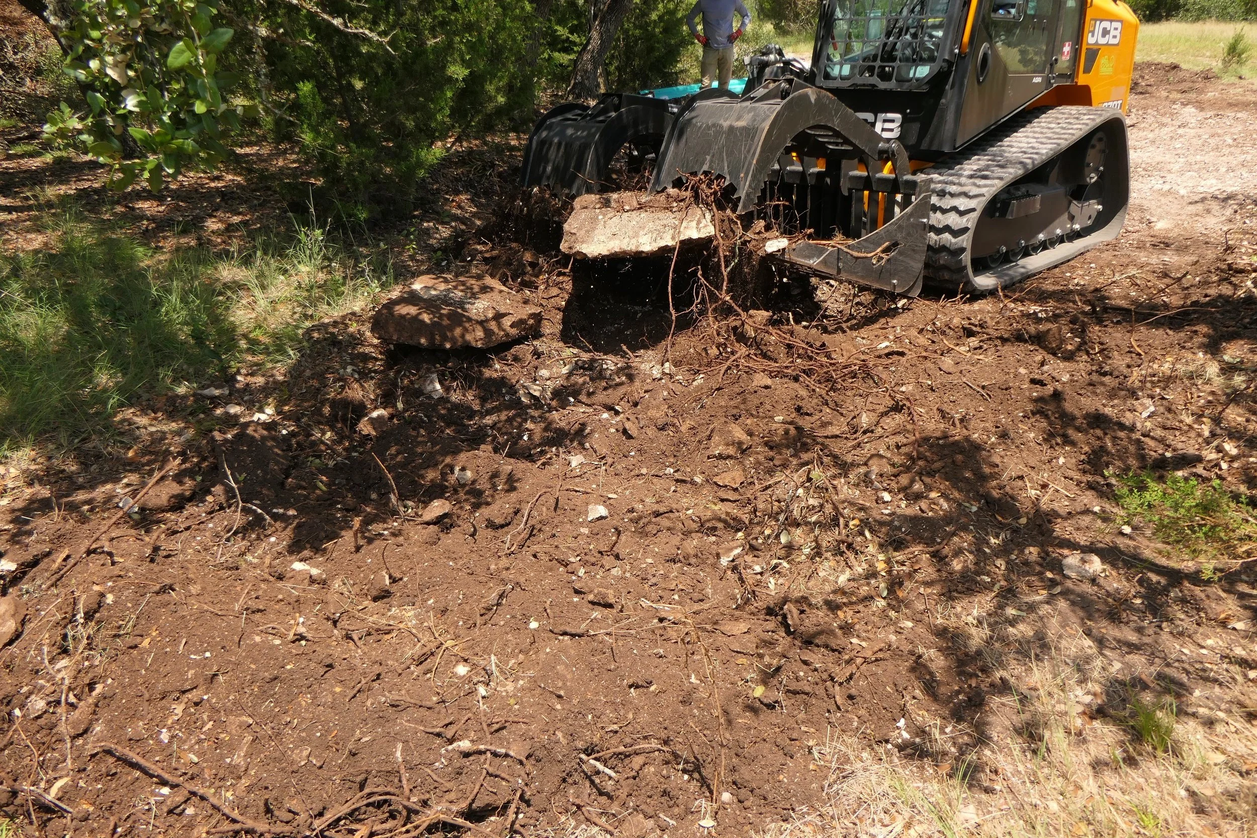 A small excavator removing dirt and roots from a soil bed outdoors, with a person standing nearby.