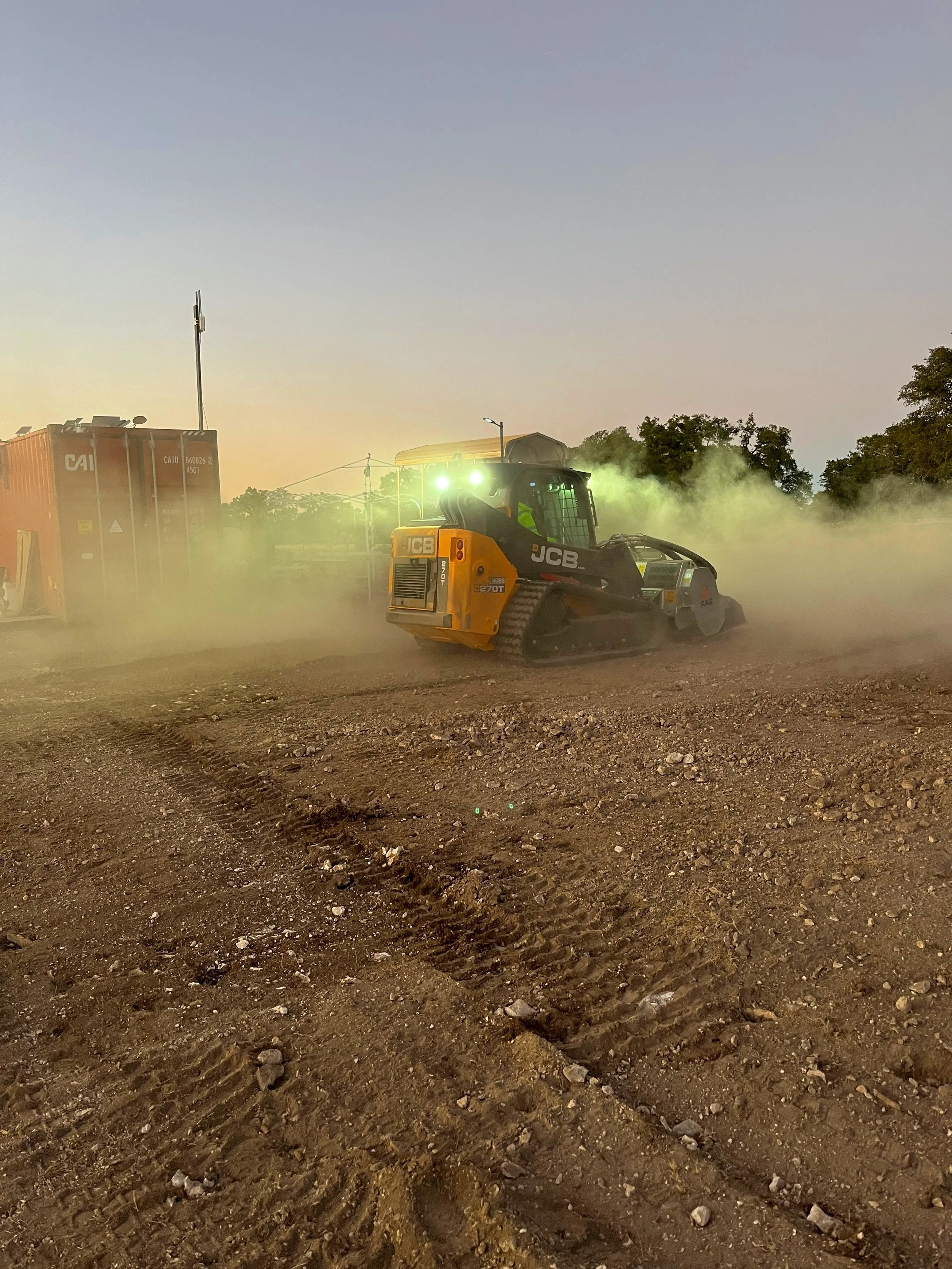 A construction site at dusk with a JCB compact track loader moving dirt, creating dust and tire tracks on the ground, with some trees and construction equipment visible in the background.