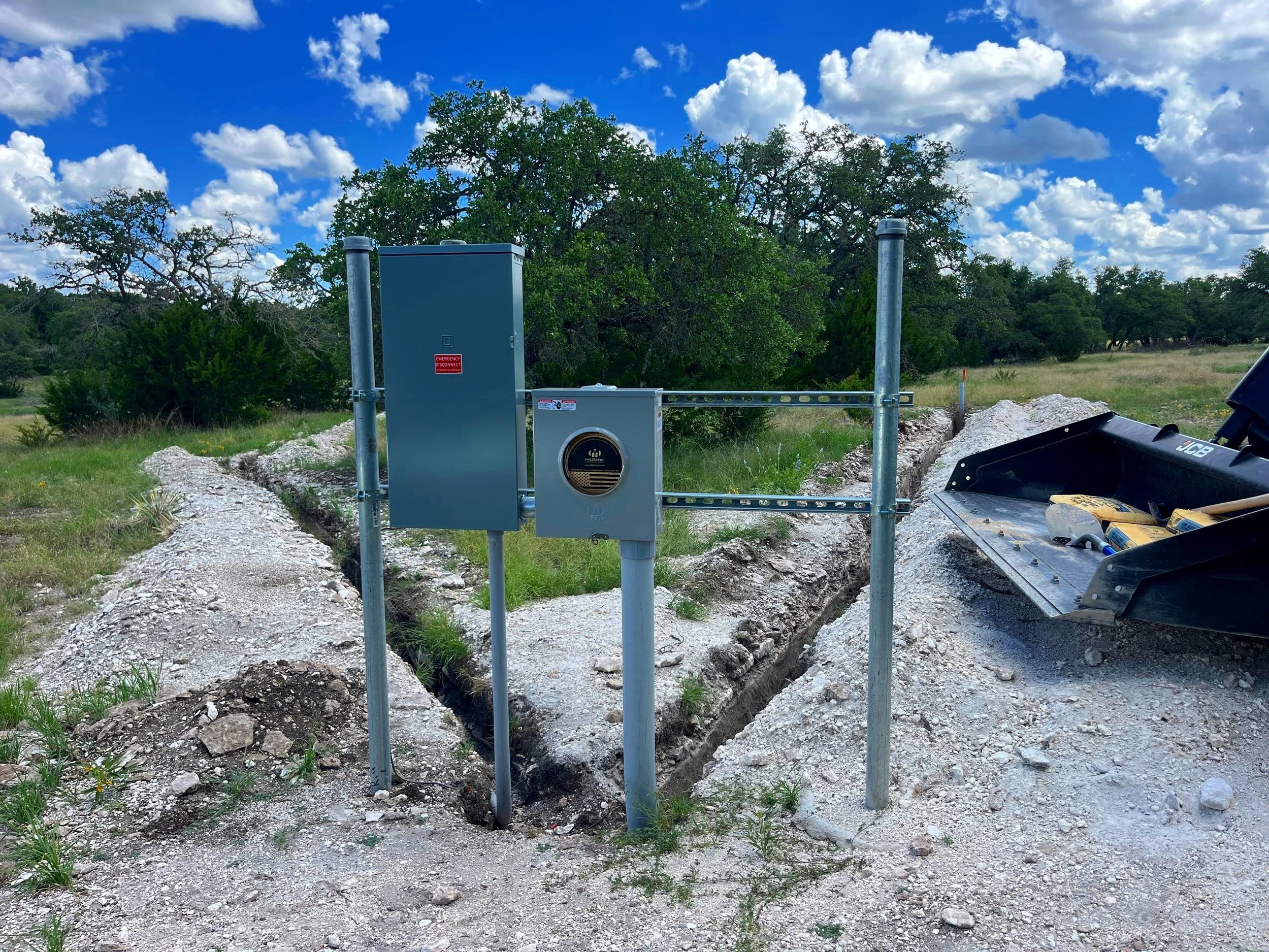Electrical infrastructure with a metal box and conduit on a construction site with a excavator, green grass, trees, and a blue sky with clouds in the background.