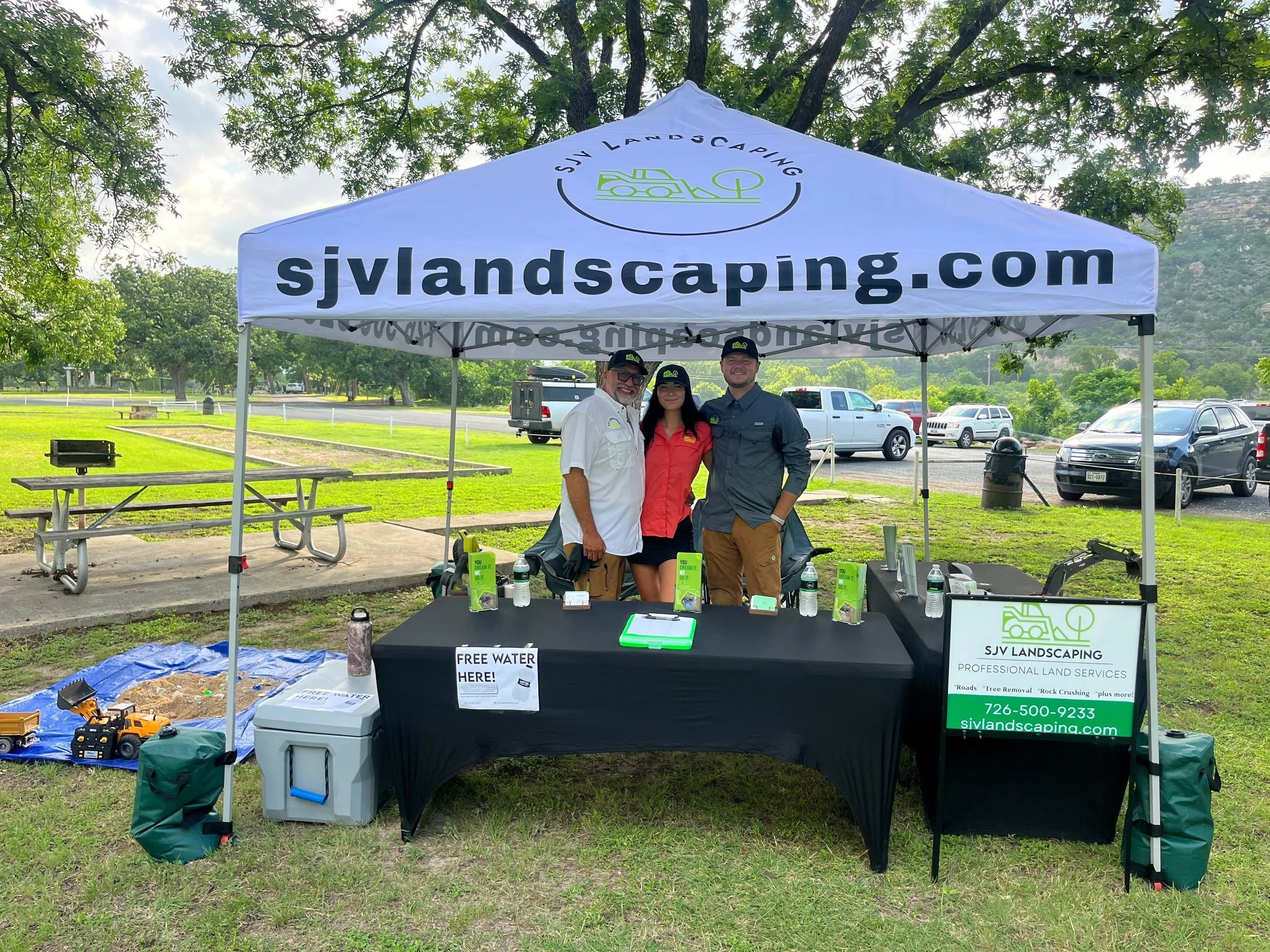 Three people standing under a white tent with the website 'sjvlandscaping.com' printed on it. The tent belongs to SJV Landscaping, which offers professional land services. The table underneath the tent has brochures, bottled water, and a sign that re