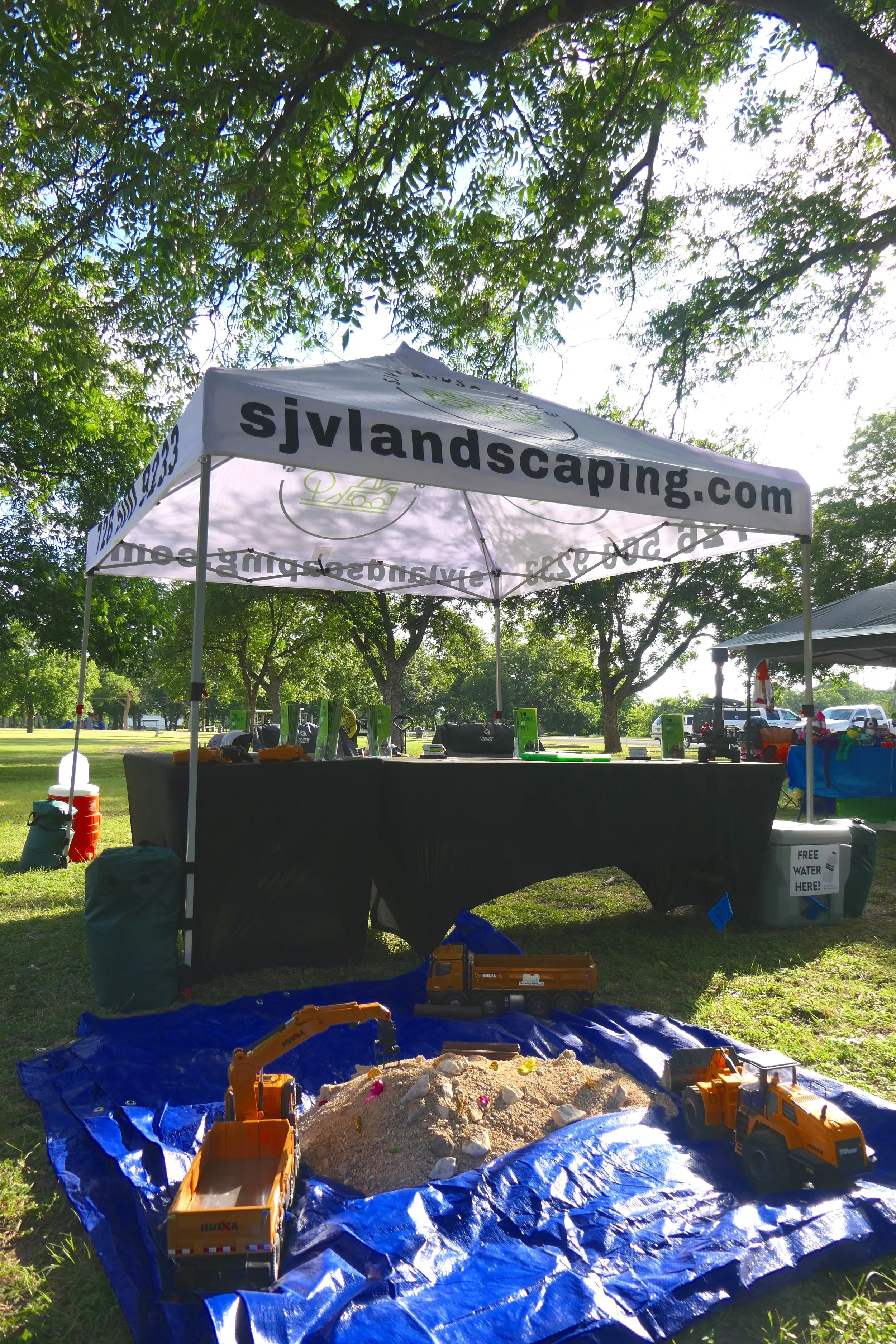 Outdoor landscaping booth with a canopy tent displaying the website sjvlandscaping.com, set up in a park with trees and grass, featuring toy construction vehicles and a blue tarp with a sand pile.