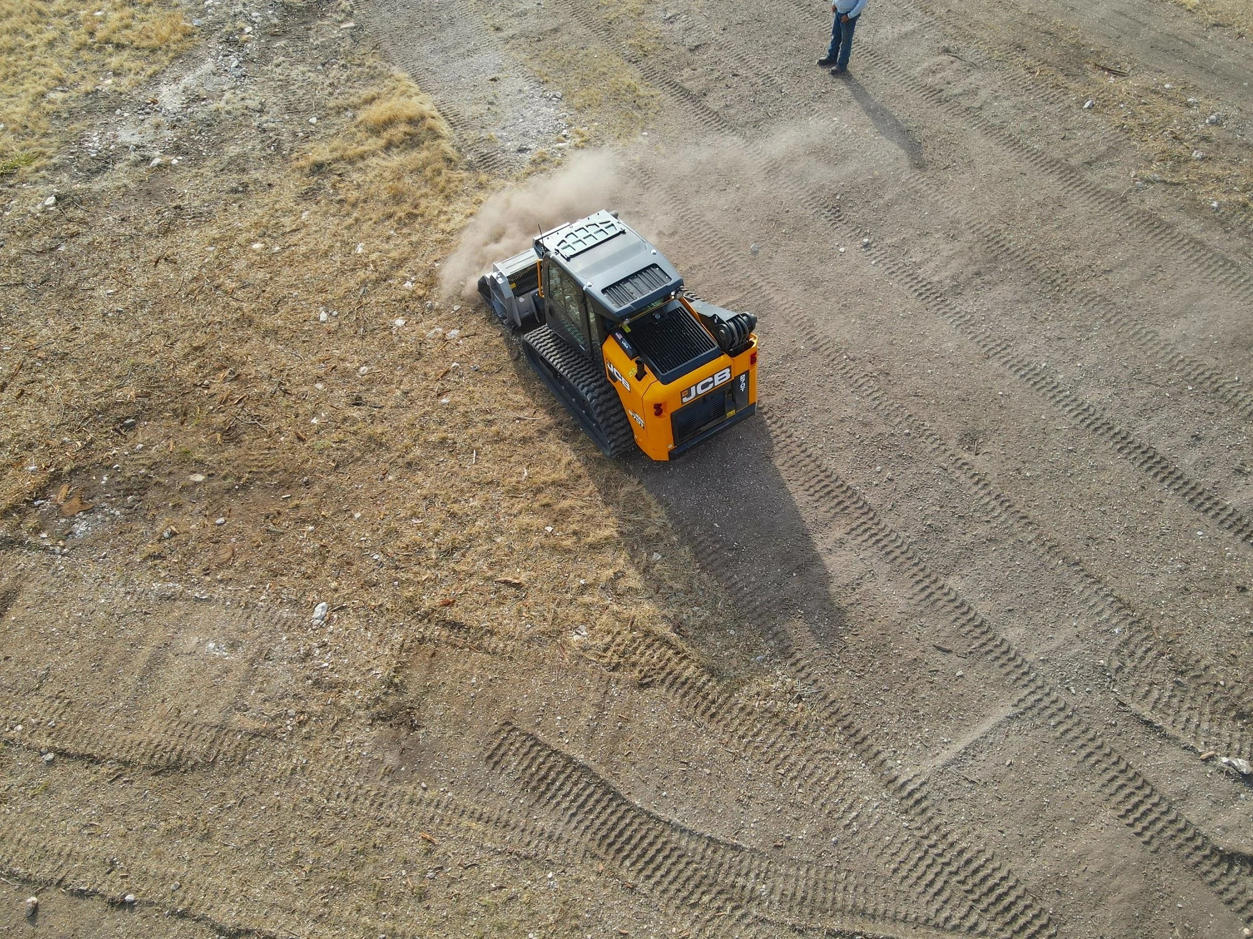A small yellow and black tracked excavator operating on dirt ground, with dust rising behind it and a man standing nearby.