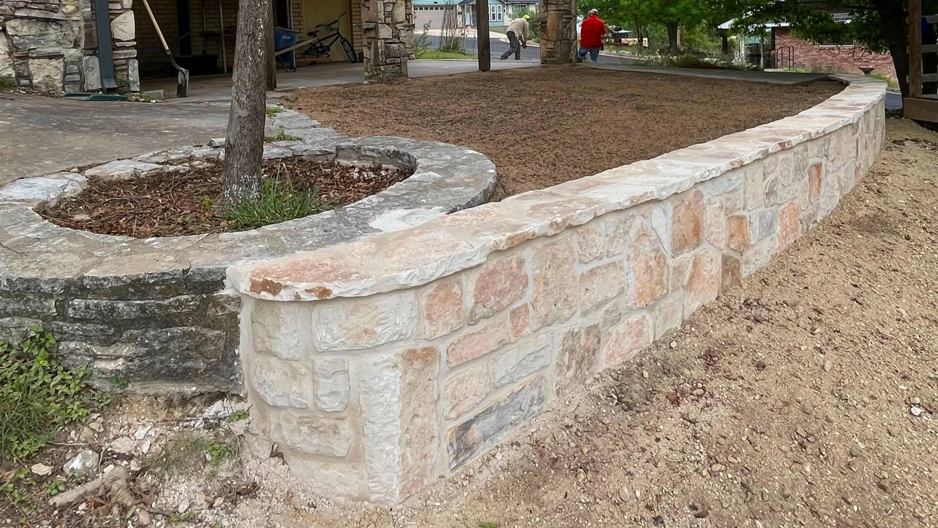 A stone brick and mortar raised garden bed with a small tree inside, filled with soil, situated on a dirt surface in an outdoor area.