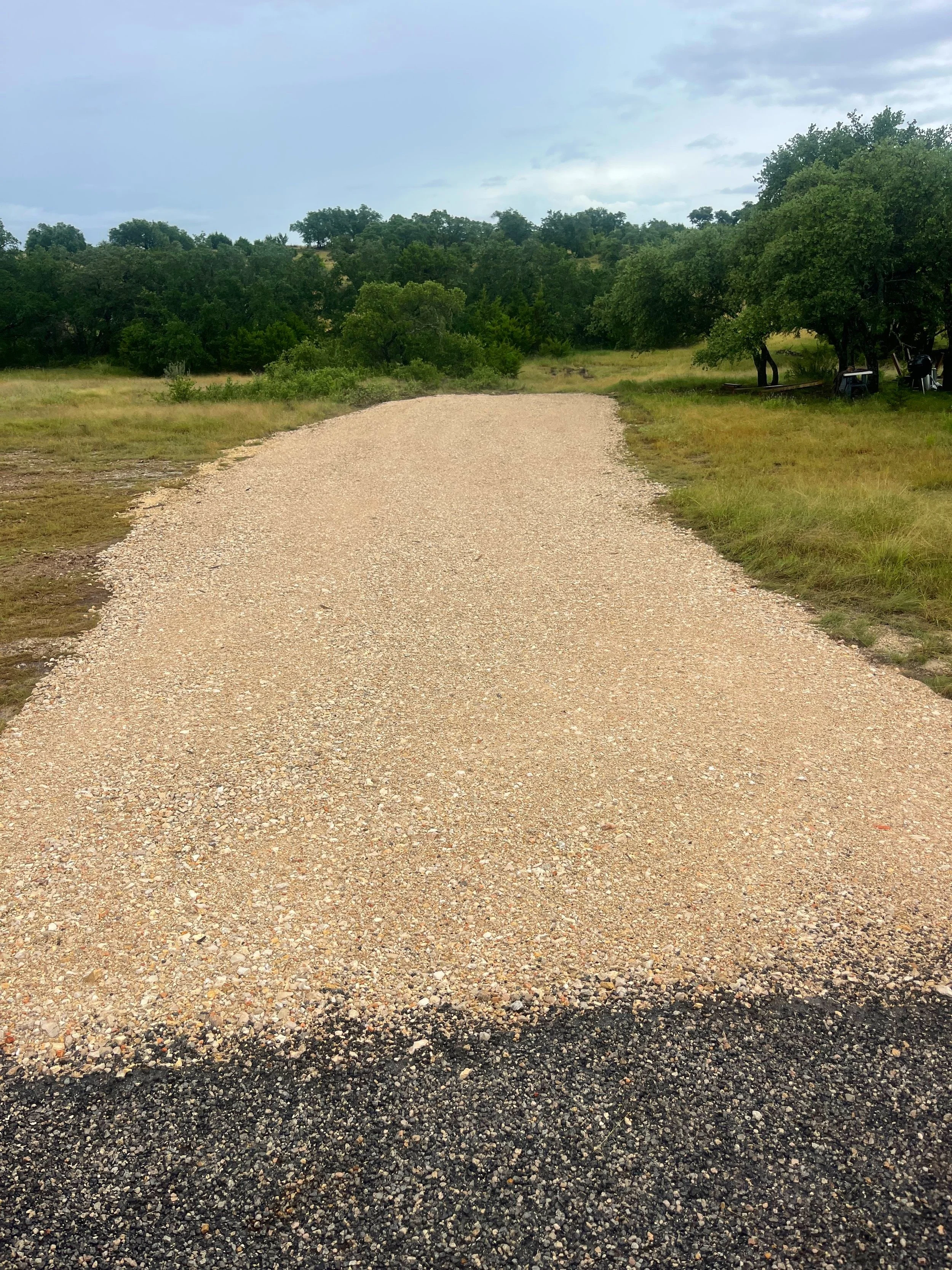 Gravel path winding through a grassy field with trees and hills in the background under a partly cloudy sky.