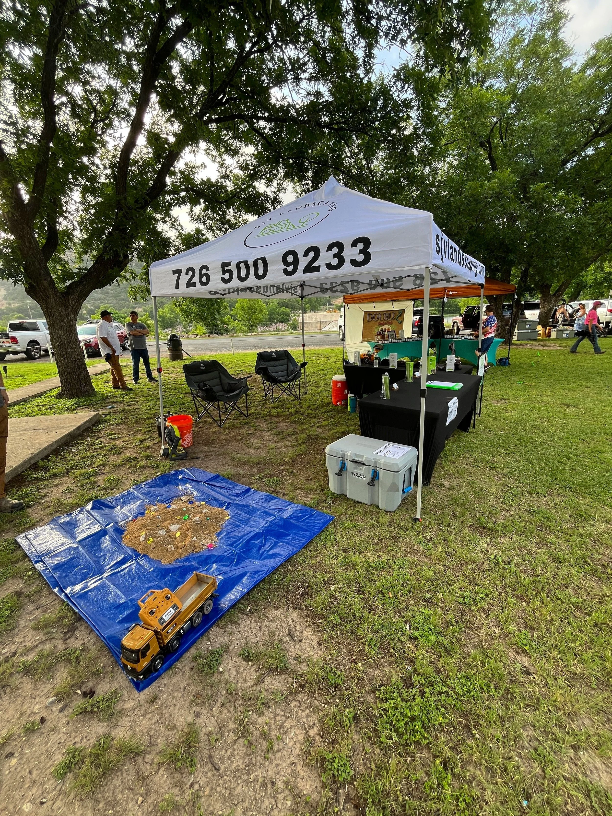 Outdoor marketplace stall with a white canopy tent displaying a phone number, with seating for three under the tent, a small table with supplies, a plastic storage container, a sandbox with toys, and toy construction vehicle, all under trees on grass