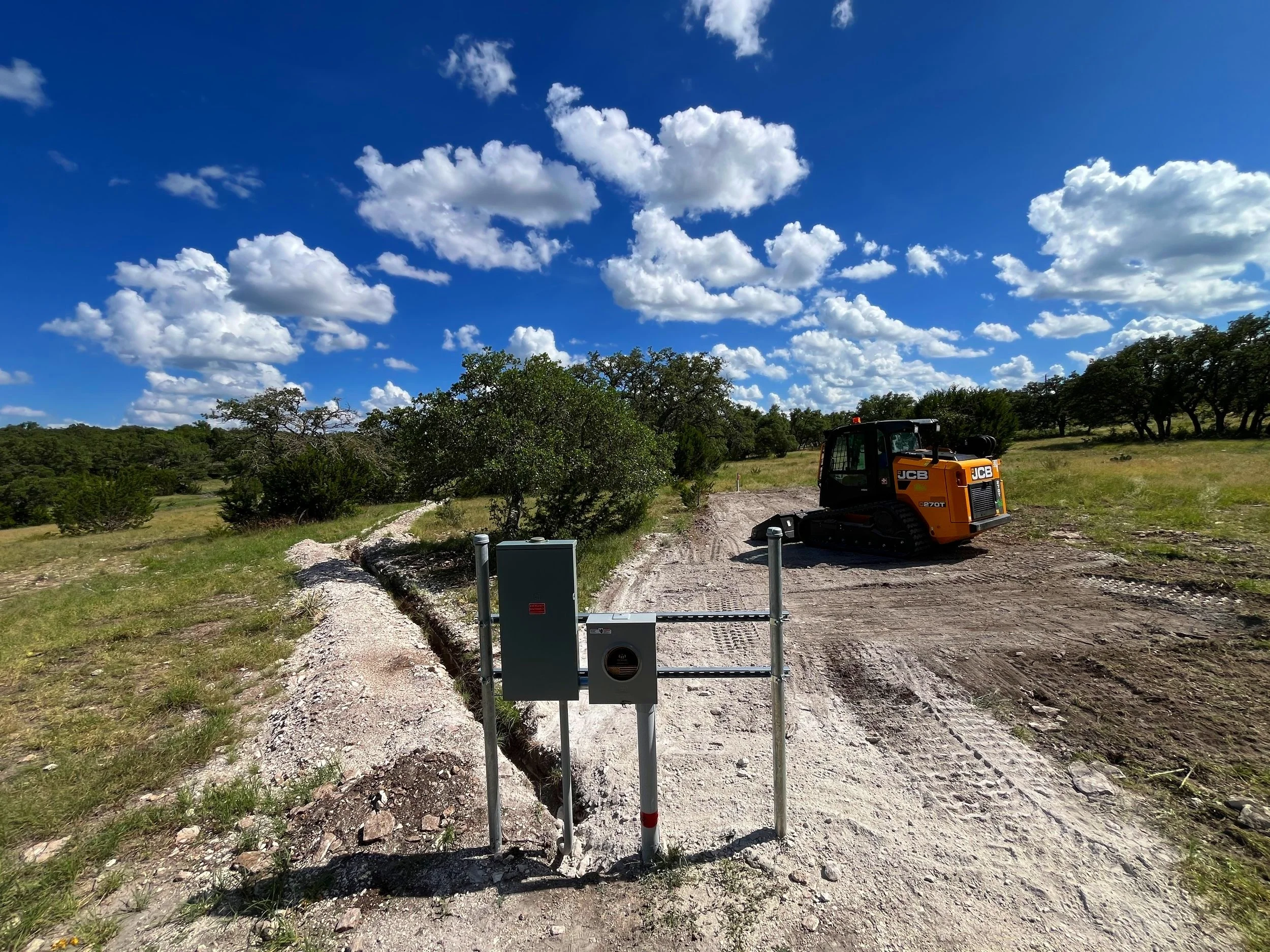 Construction site on a dirt road under a bright blue sky with scattered clouds, featuring a small orange tracked construction vehicle and utility boxes near the road.