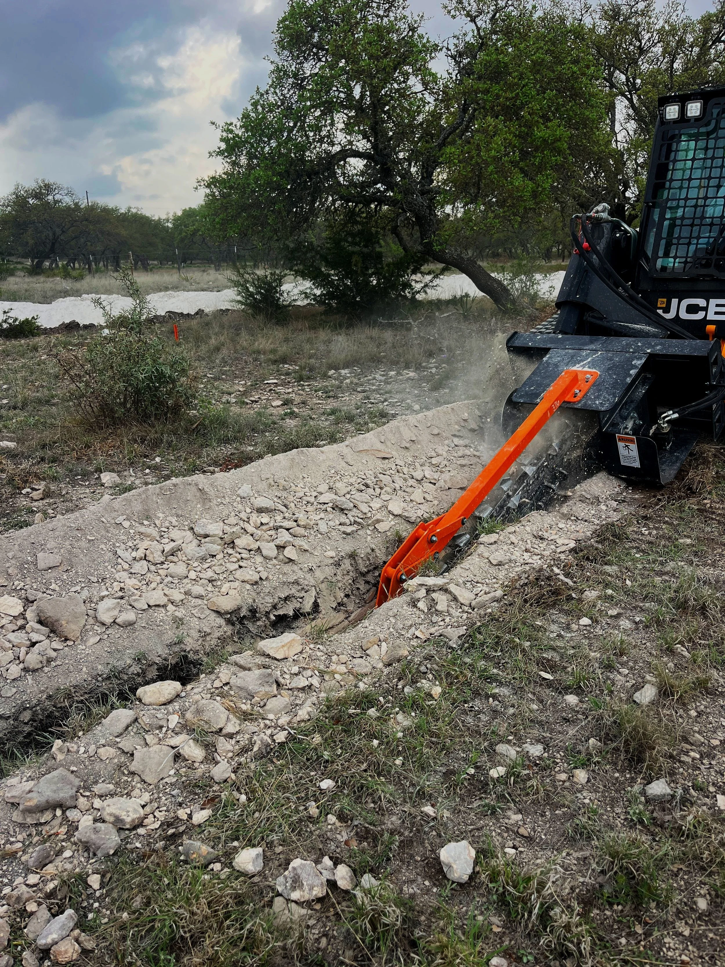 A backhoe digging a trench in a grassy field with trees and cloudy sky in the background.