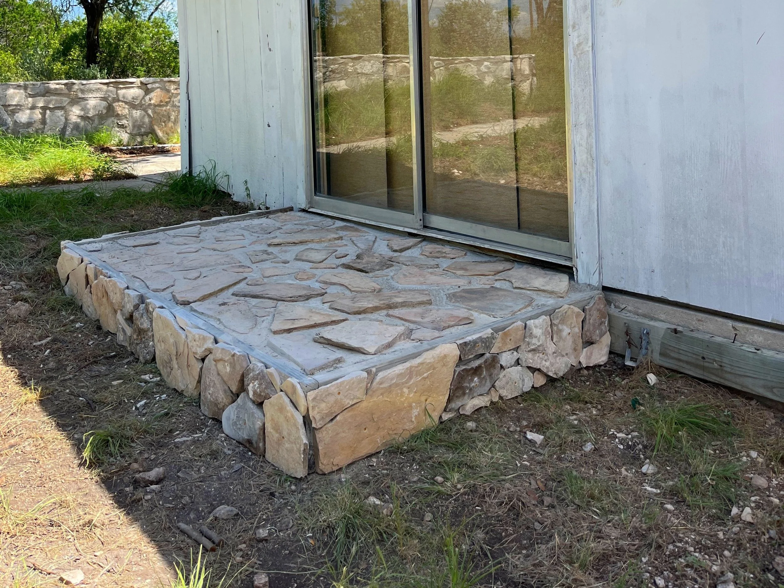 A newly constructed stone step in front of a sliding glass door on a house, with exterior siding and a stone retaining wall in the background.