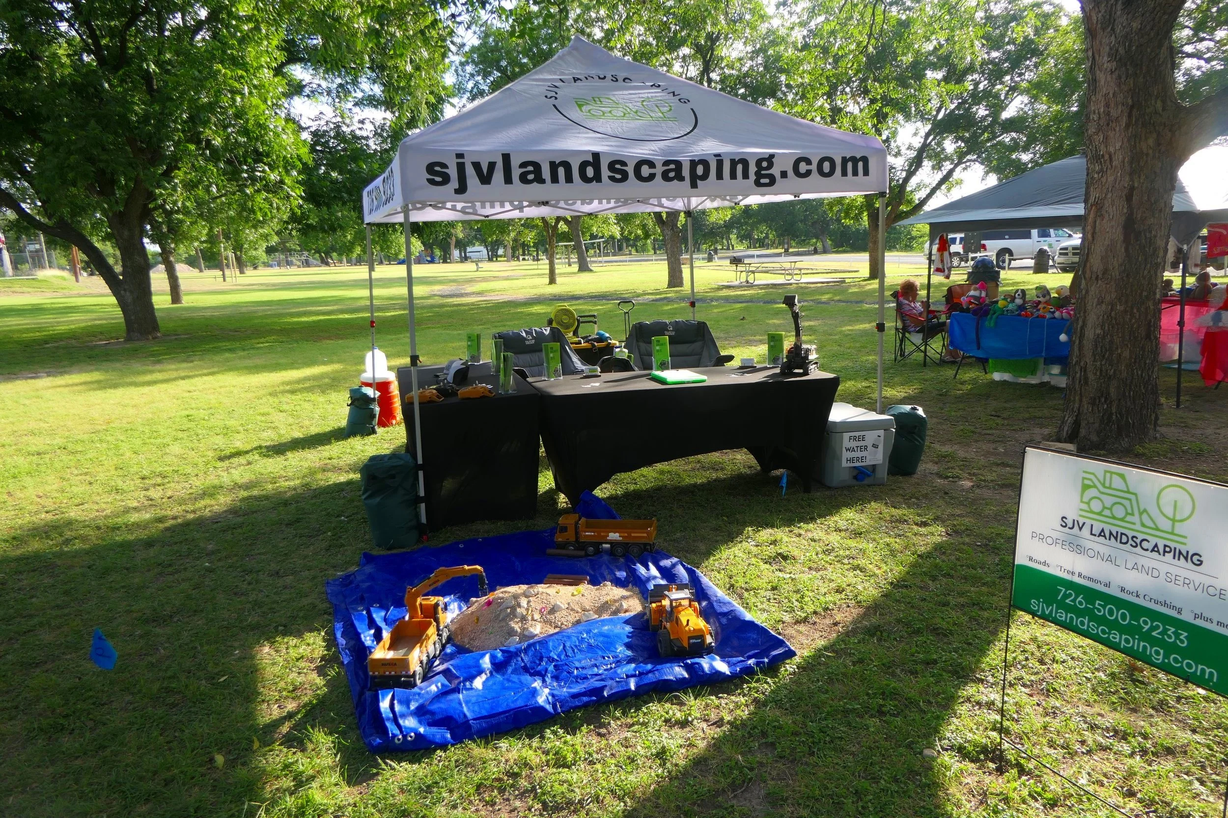 An outdoor landscaping company booth set up in a park with a canopy tent displaying the company name 'sjvlandscaping.com'. The booth has tools and promotional material on a black table, and a small sand and gravel display with toy dump trucks on a bl