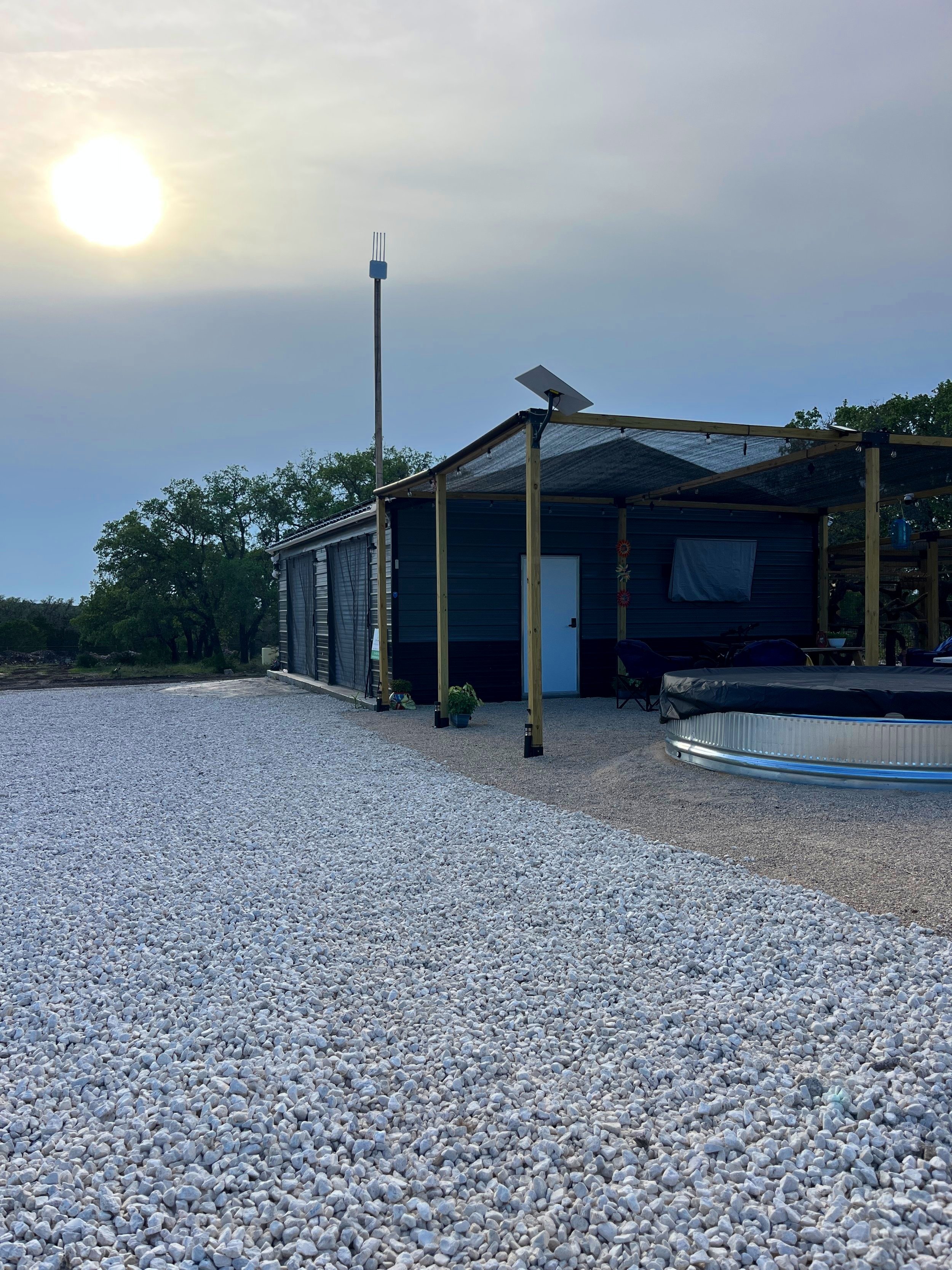 A house with a gravel driveway, small plants outside, and a pergola covering a seating area. The sun is low in the sky.