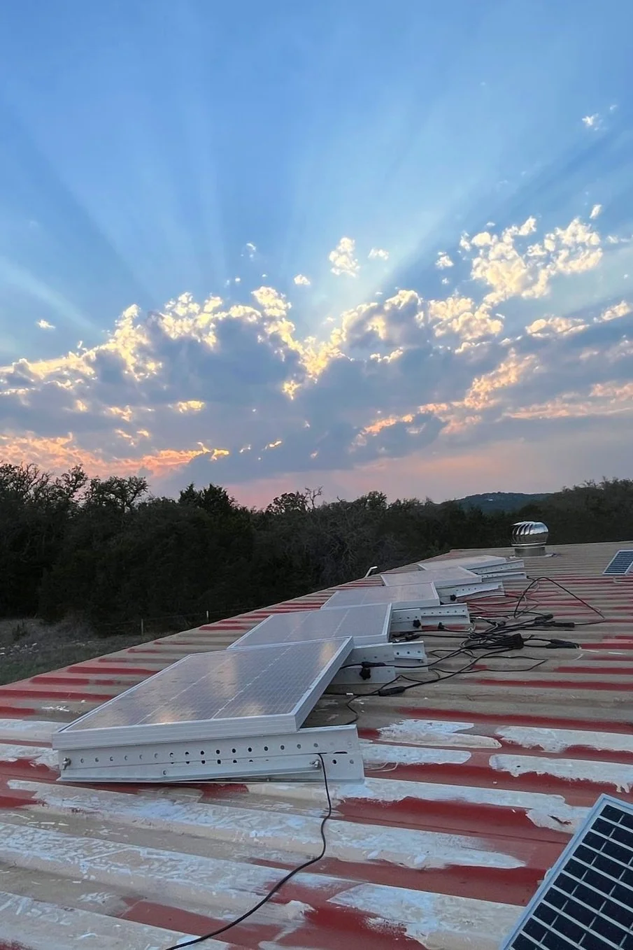 Solar panels installed on a rusty metal roof at sunset, with a partly cloudy sky and sun rays in the background.