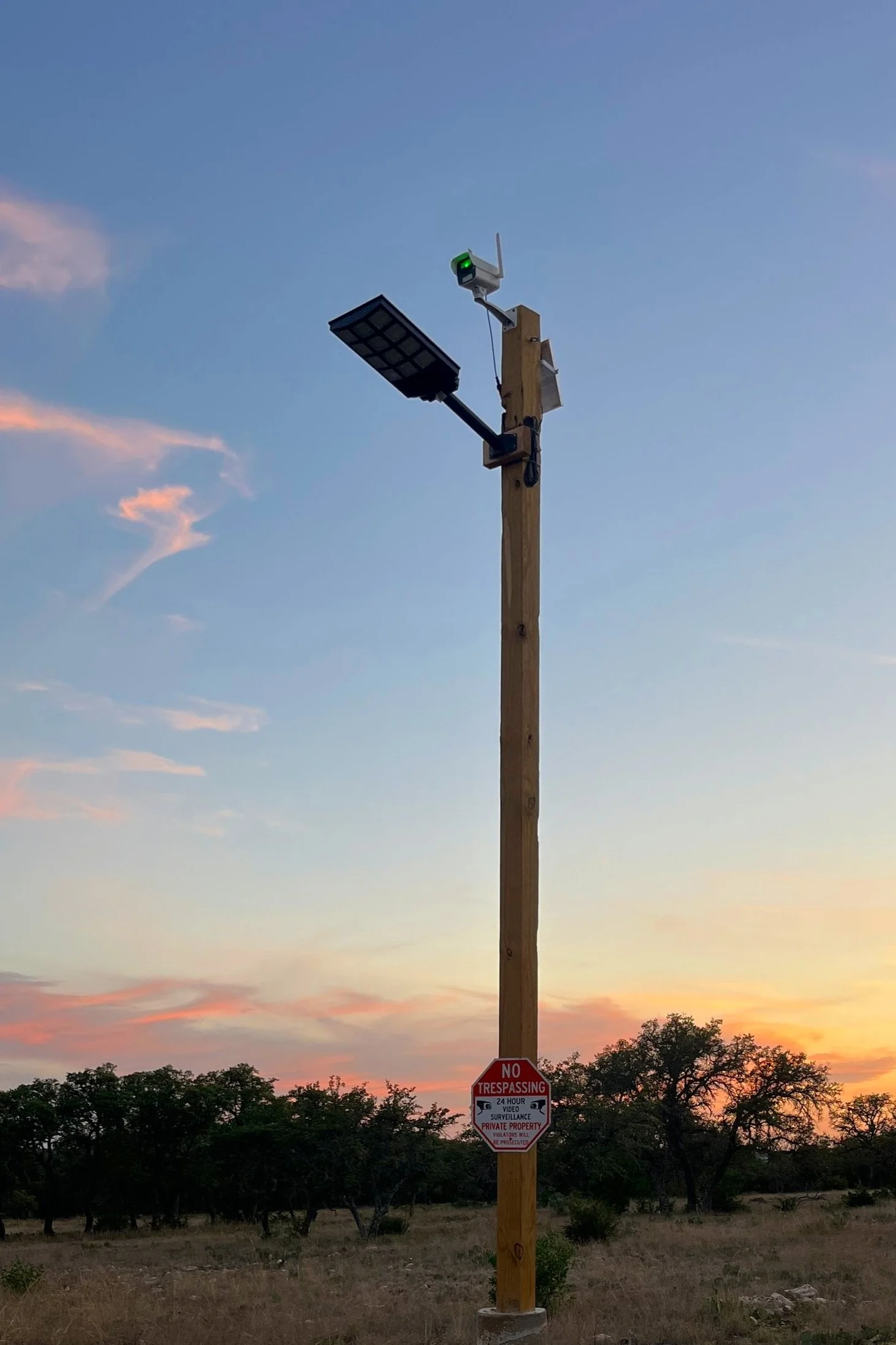 A wooden utility pole with solar panels, a camera, and a streetlight attached. There is a red 'No Trespassing' sign on the pole, set against a dusk sky with trees in the background.