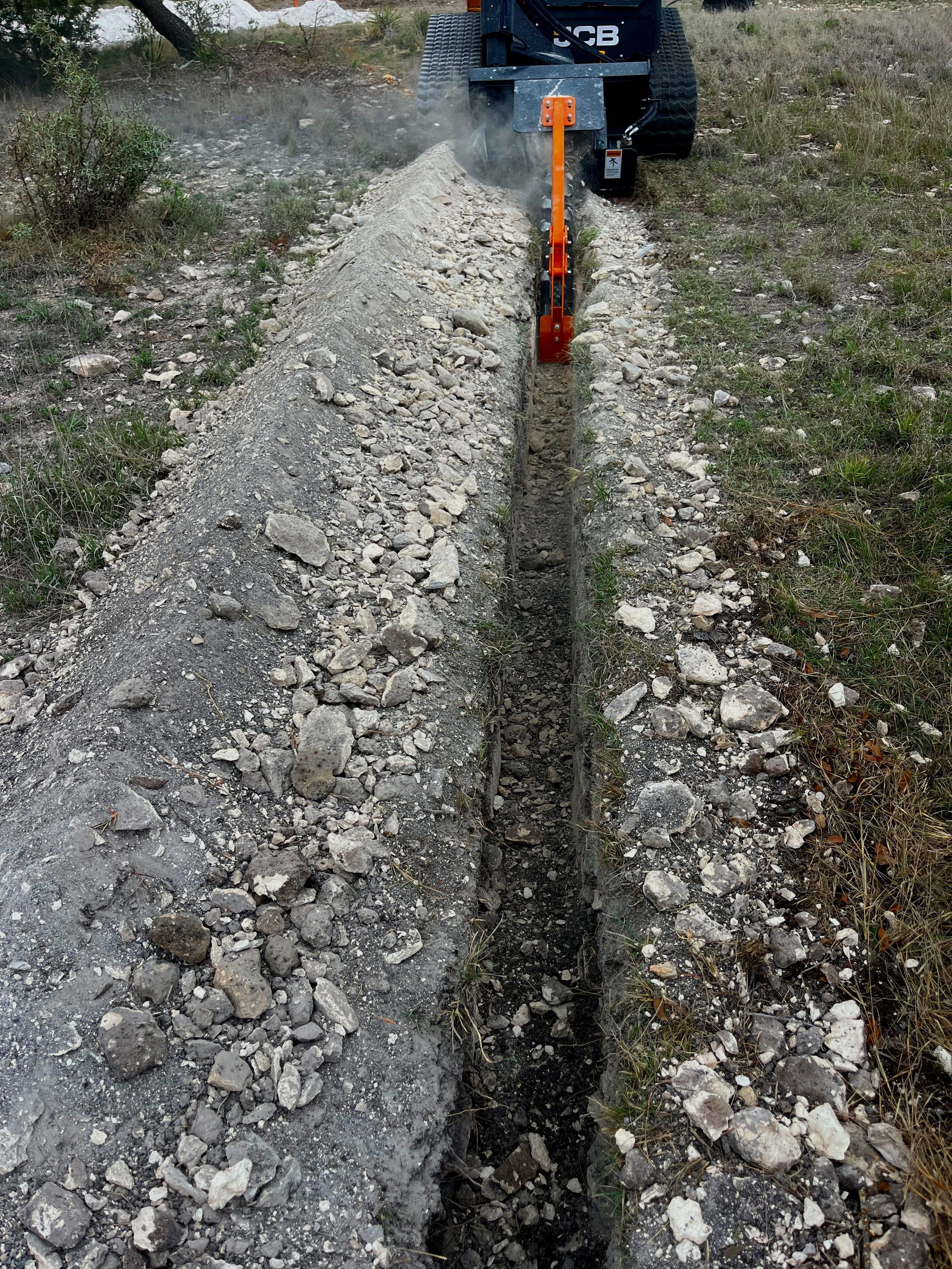 A small excavator digging a trench in a dirt field with grass and shrubs on either side.