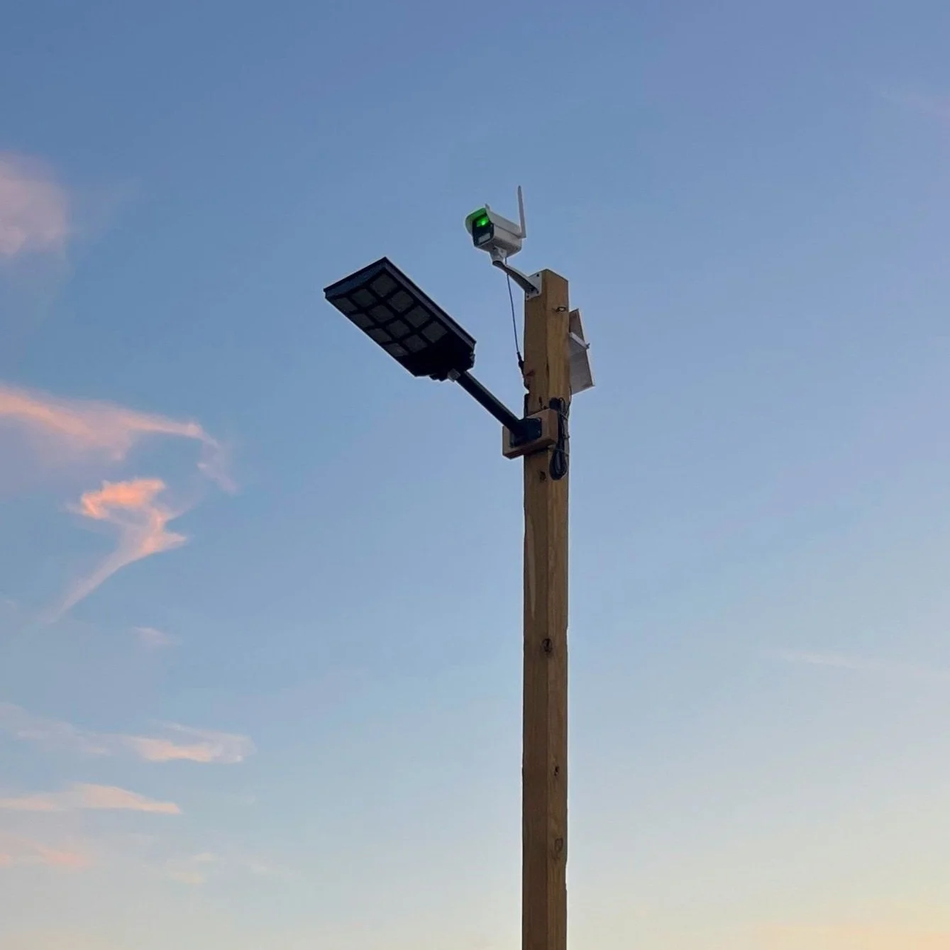 A wooden utility pole with a green traffic light attached, topped with a security camera, a solar panel, and other small electronic devices against a blue sky with pink clouds.