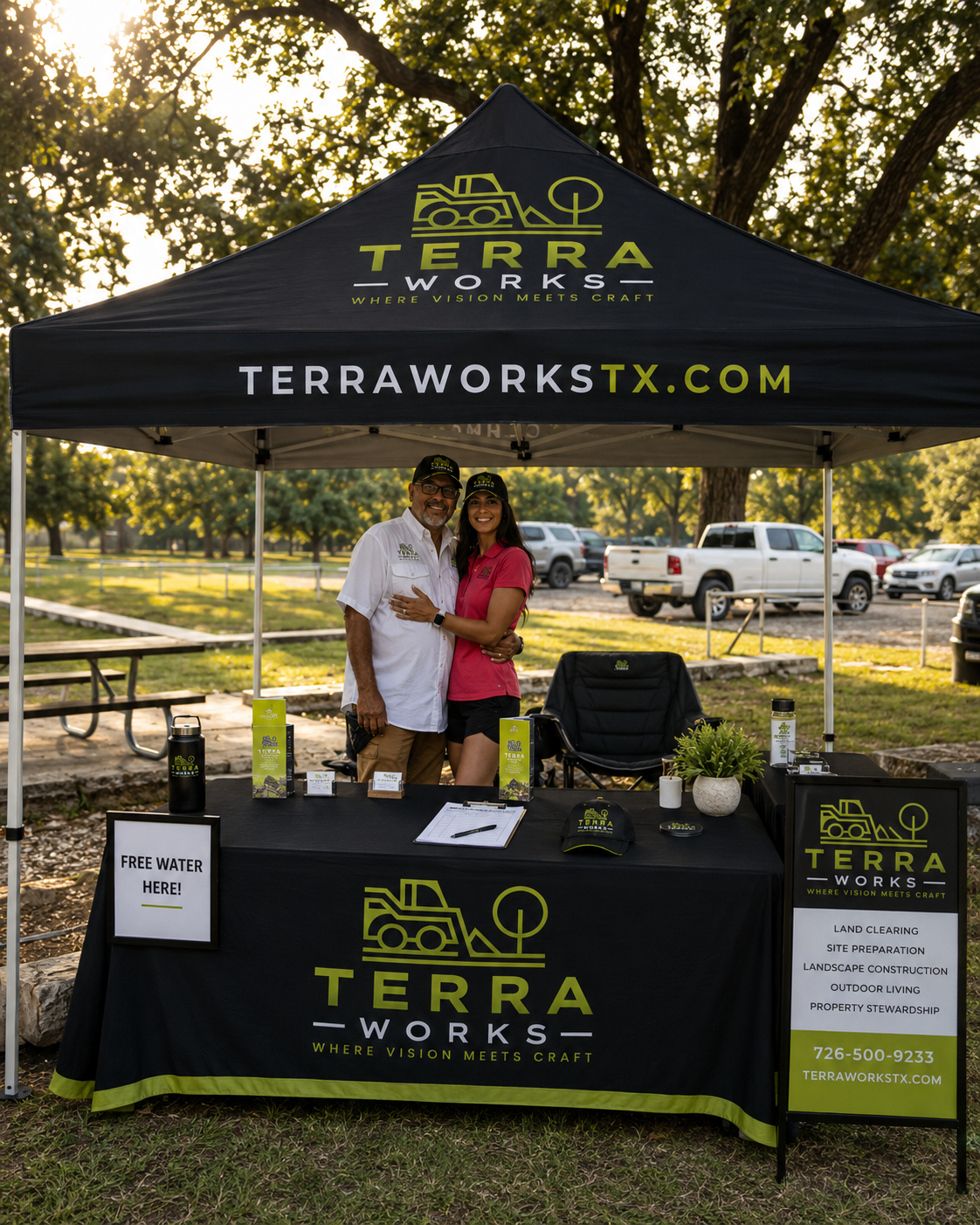 Two people standing under a black Terra Works tent at an outdoor event, smiling and embracing. The tent displays the company logo and website, with a table set up with promotional materials, a water bottle, and a sign offering free water. Trees and parked cars are visible in the background.