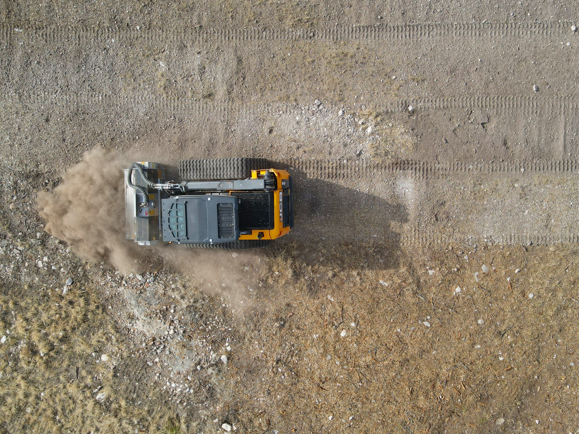An aerial view of a yellow and gray compact soil compactor rolling over dirt, with dust and tire tracks visible on the ground.