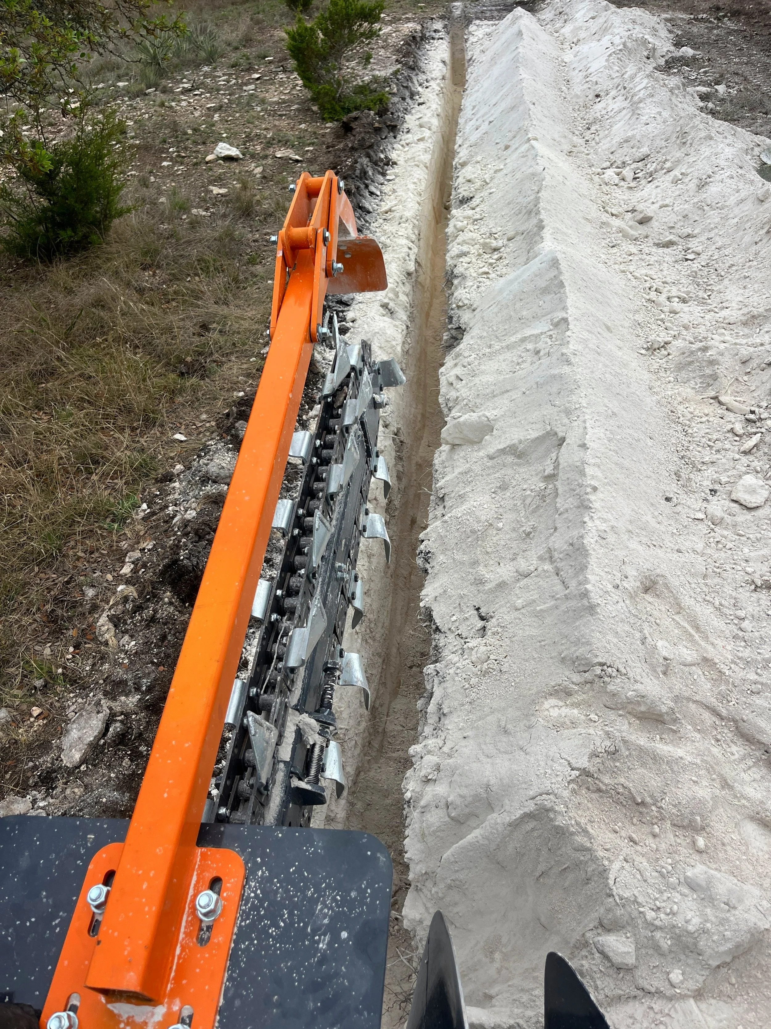 Close-up of an orange and metallic trencher blade cutting into a trench, with a large mound of dirt on the right side.