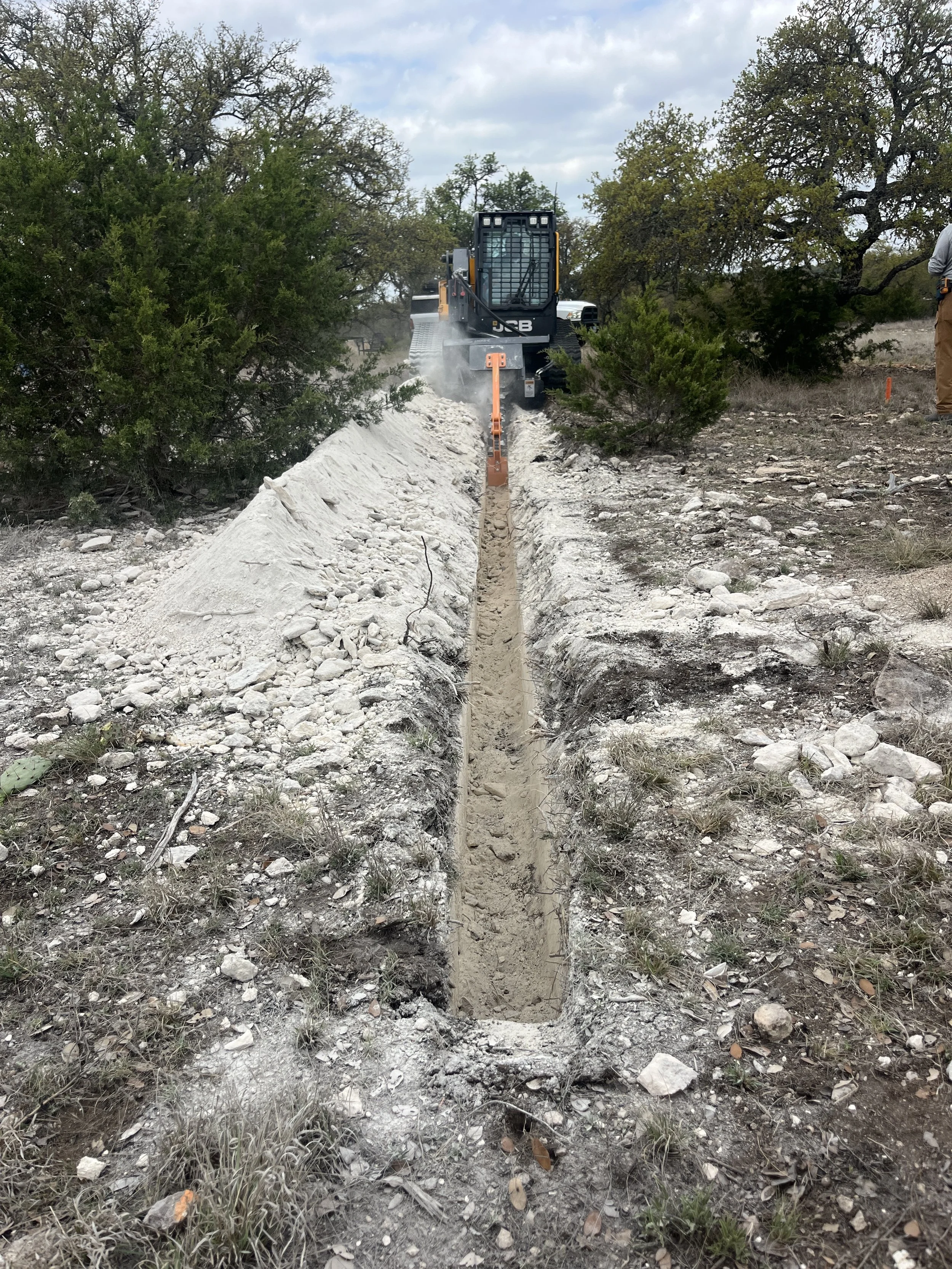 Construction site with a narrow trench being dug in the ground, with a machine working at the far end of the trench amidst trees and a cloudy sky.