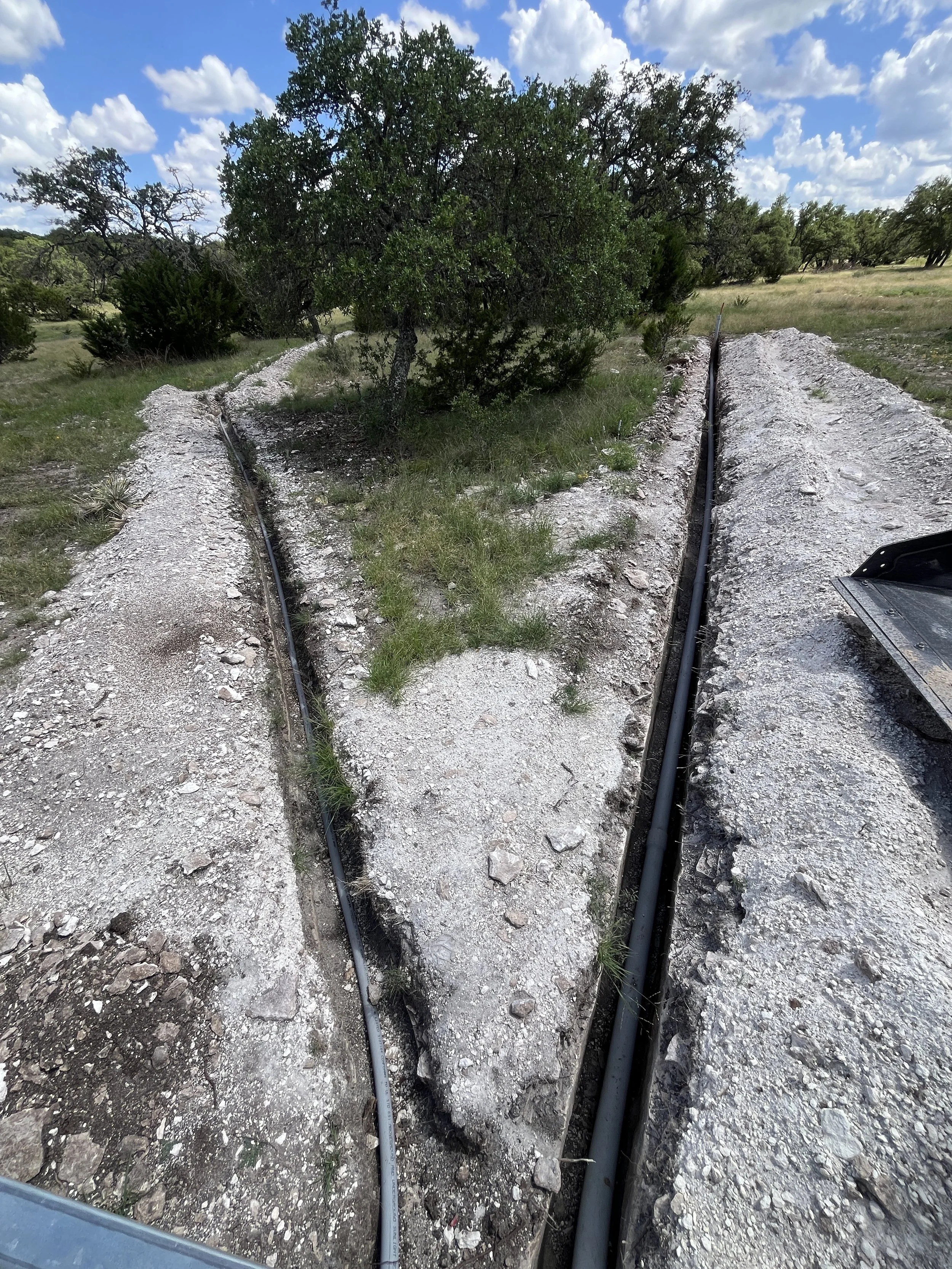 A dirt road with exposed electrical conduit pipes running along each side in a rural landscape with grass, trees, and blue sky with clouds.