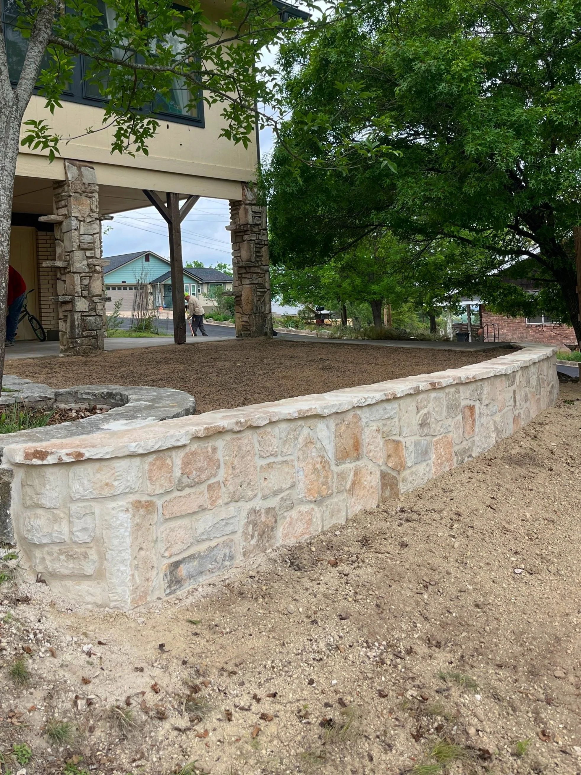 A newly built stone retaining wall with a dirt top surface, adjacent to a house with a porch supported by stone columns, with trees and houses in the background.