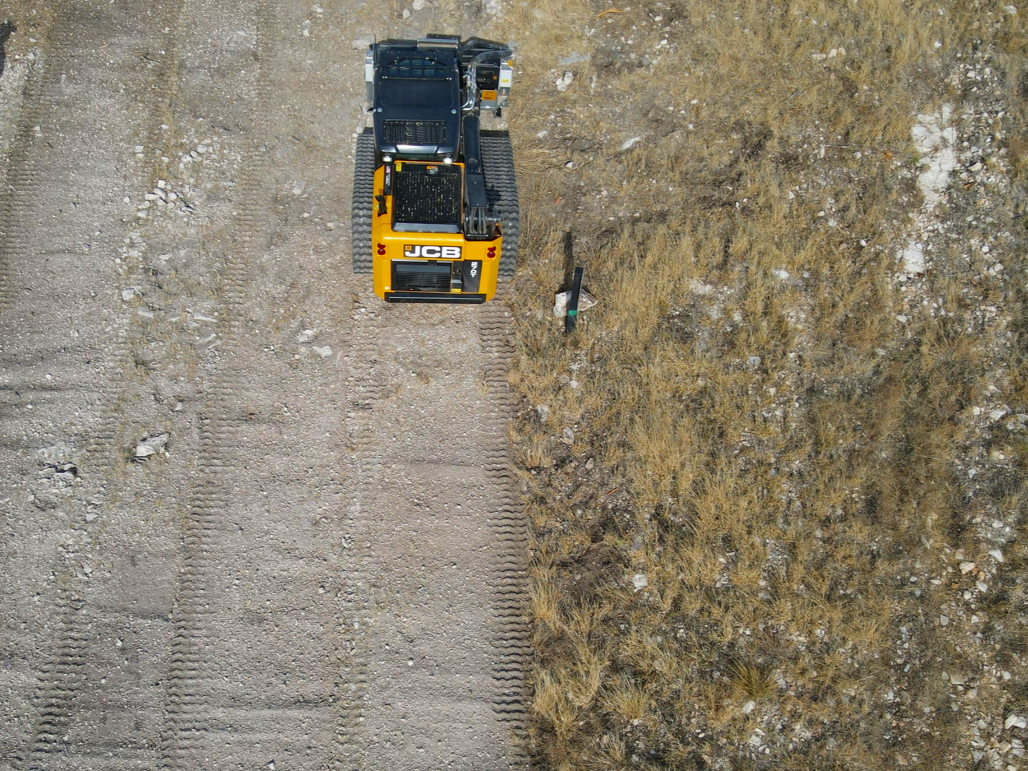 A yellow and black JCB excavator working on a dirt construction site with a grassy and rocky terrain beside it.