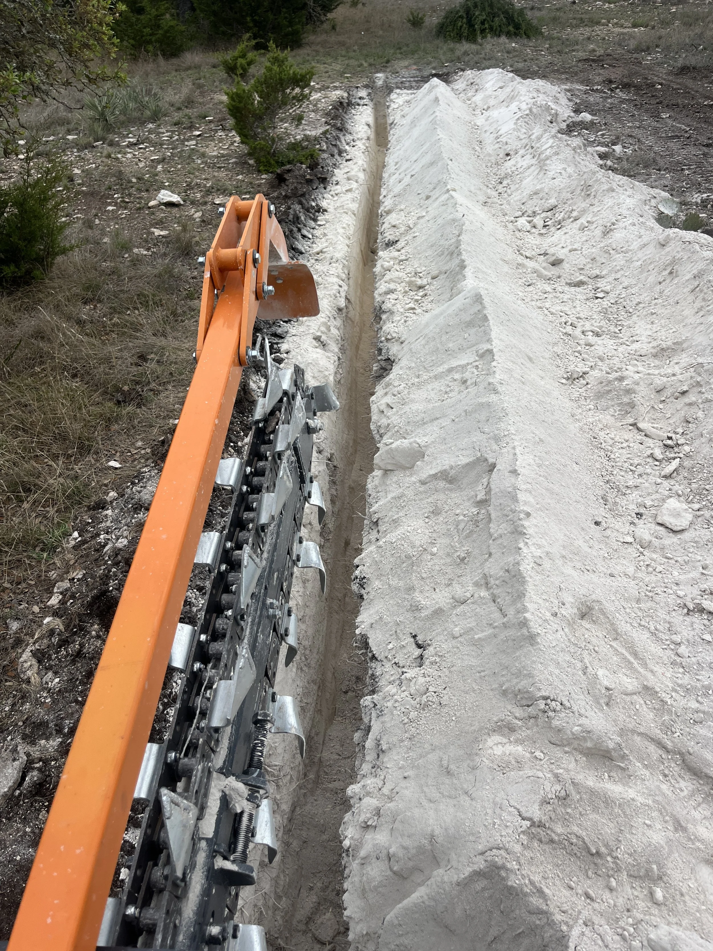 Close-up of an orange rail with a chain attached, alongside a narrow track of dirt and white powdery soil, with some greenery in the background, possibly at a construction site.
