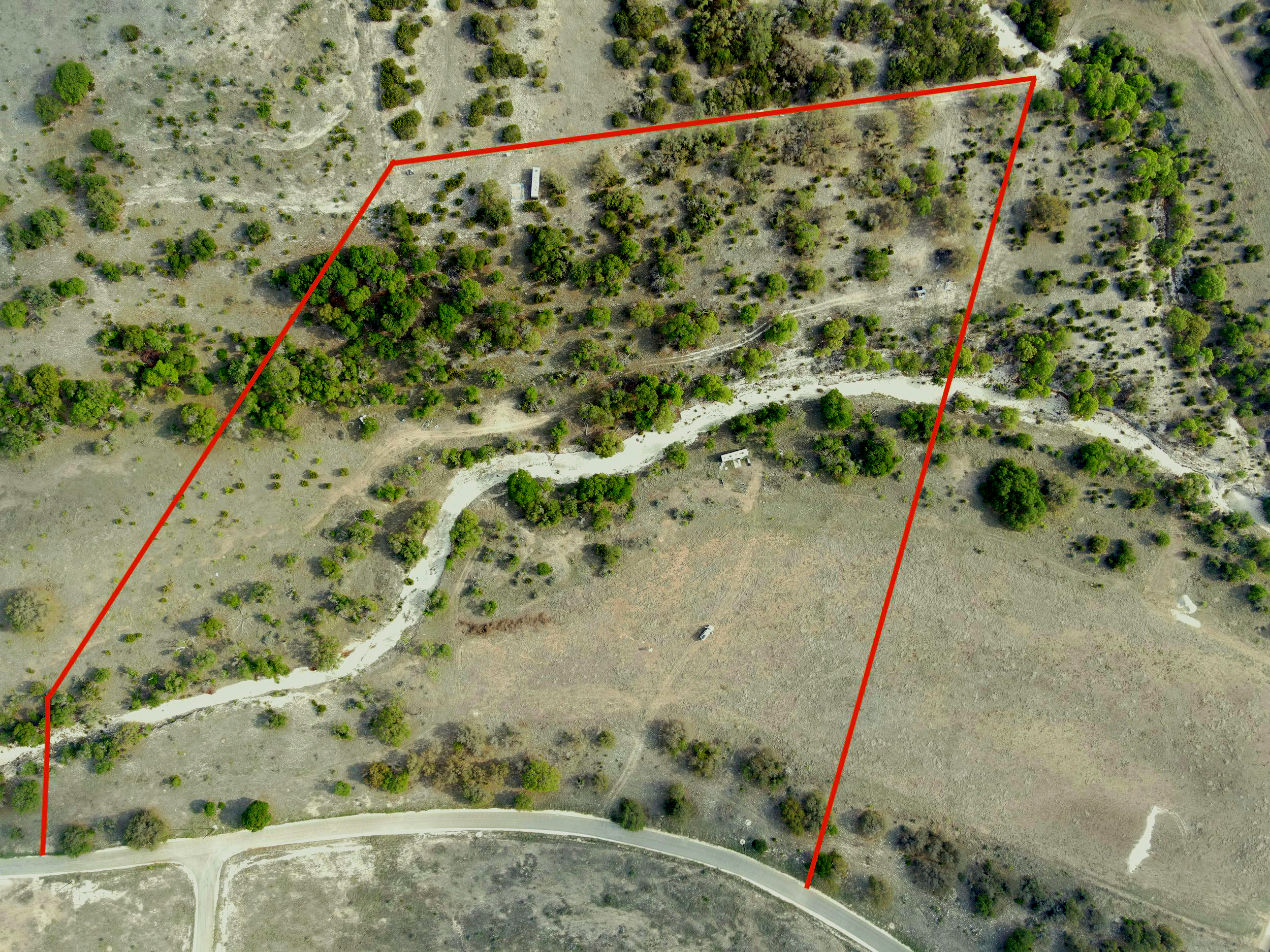 Aerial view of a plot of land with trees, dirt paths, a small building, and a curved paved road at the bottom. Red boundary lines outline the plot.