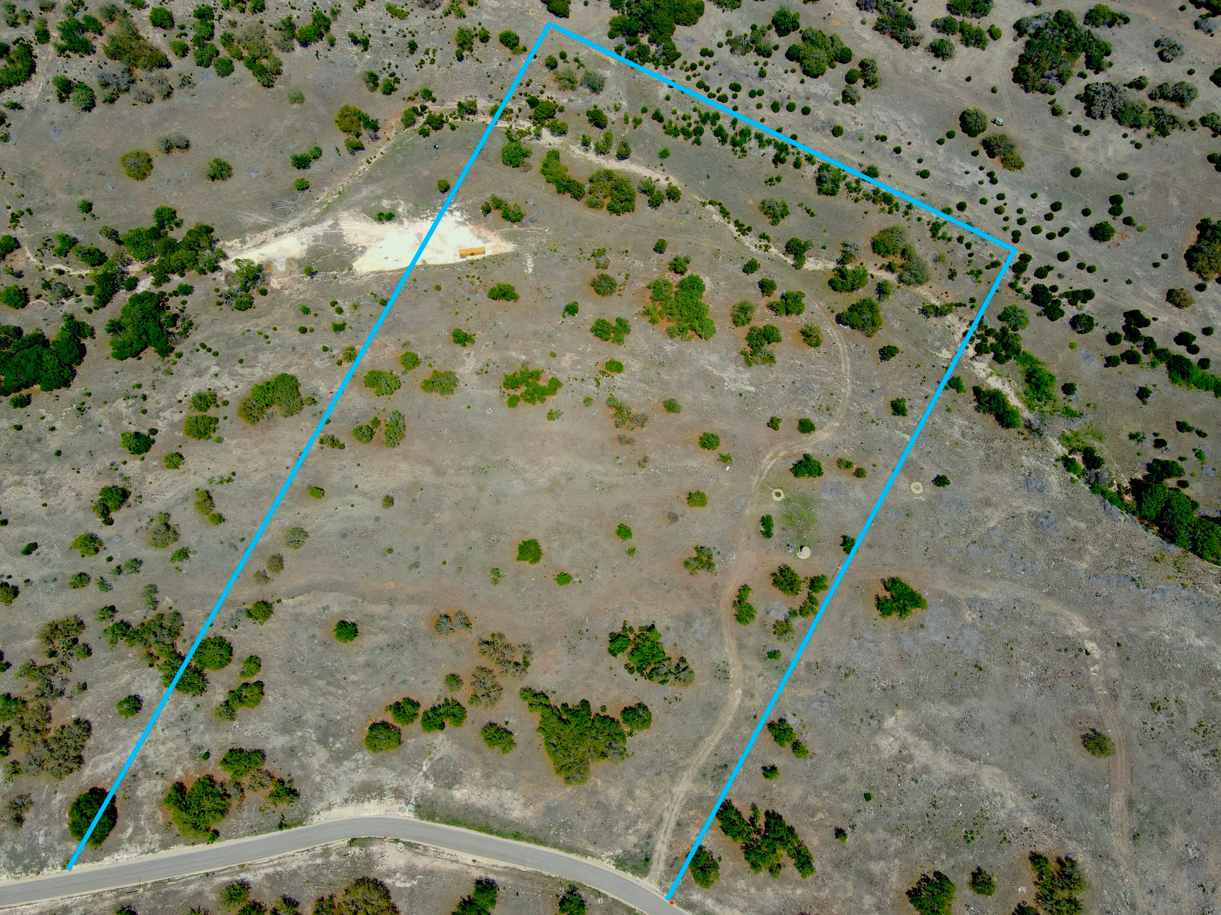 Aerial view of a large, open, grassy area enclosed by a blue boundary line, with scattered trees and a dirt pathway in the middle, near a curved road at the bottom of the image.