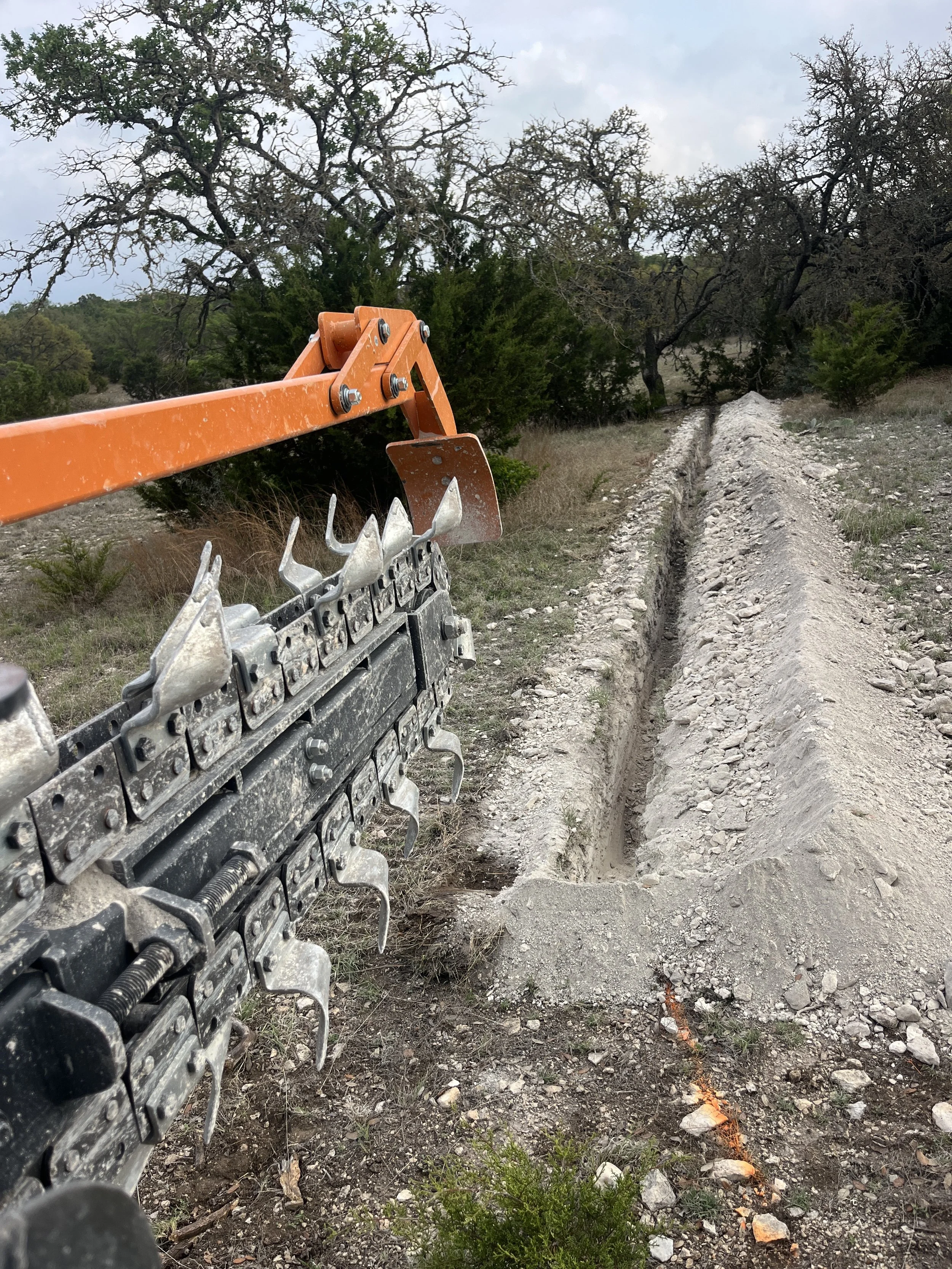 A trench being dug in a rural outdoor area with dirt piles on each side, using an orange trenching machine, with trees and cloudy sky in the background.
