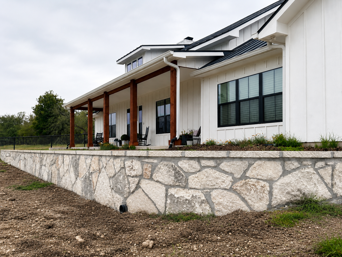 White house with black window frames, a stone foundation, and a porch supported by wood beams, with a dirt yard in front.