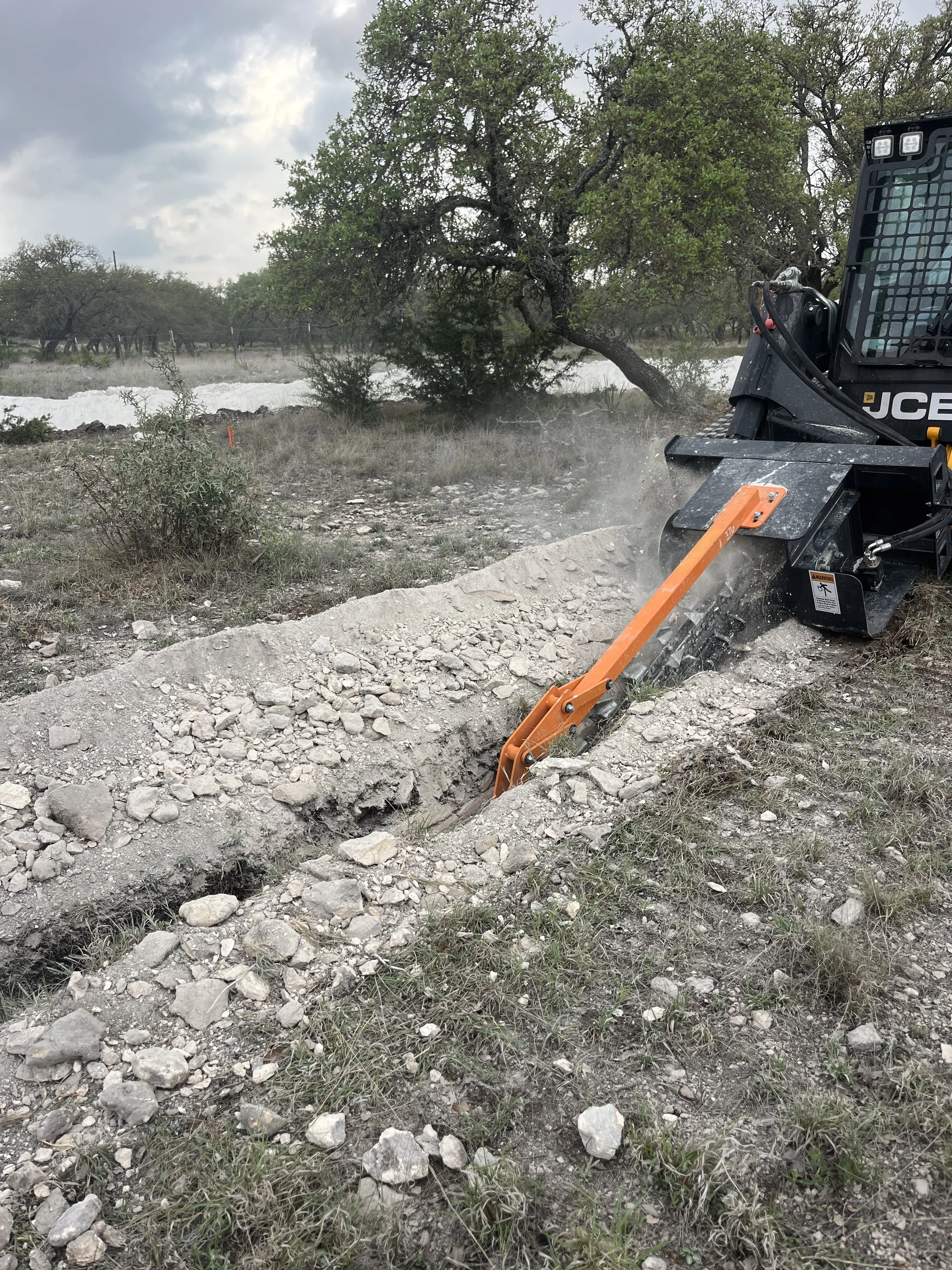 A trencher attachment digging a trench in a caliche rock filled, rural area with trees and cloudy sky in the background.