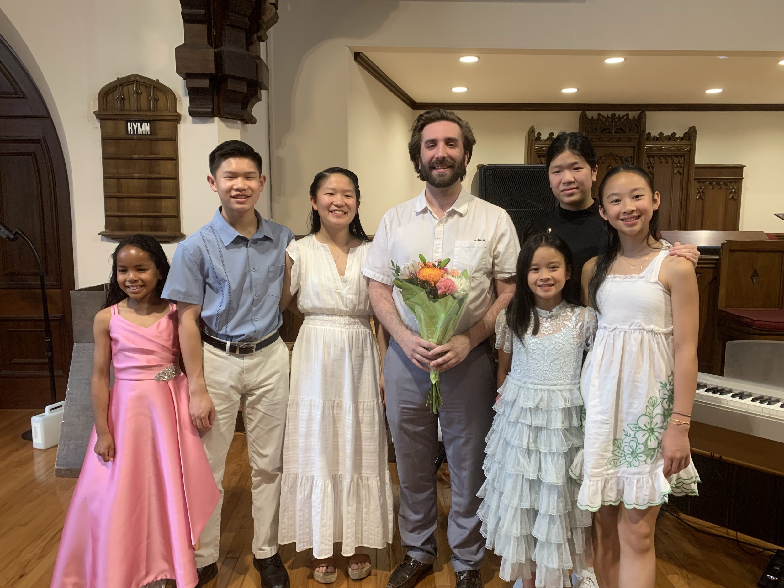 A group of seven people, including six children and one adult man, standing in a church or hall, dressed in formal or semi-formal clothing. The man is holding a bouquet of flowers and is smiling. The children are also smiling, and the background features wooden decor and a piano.