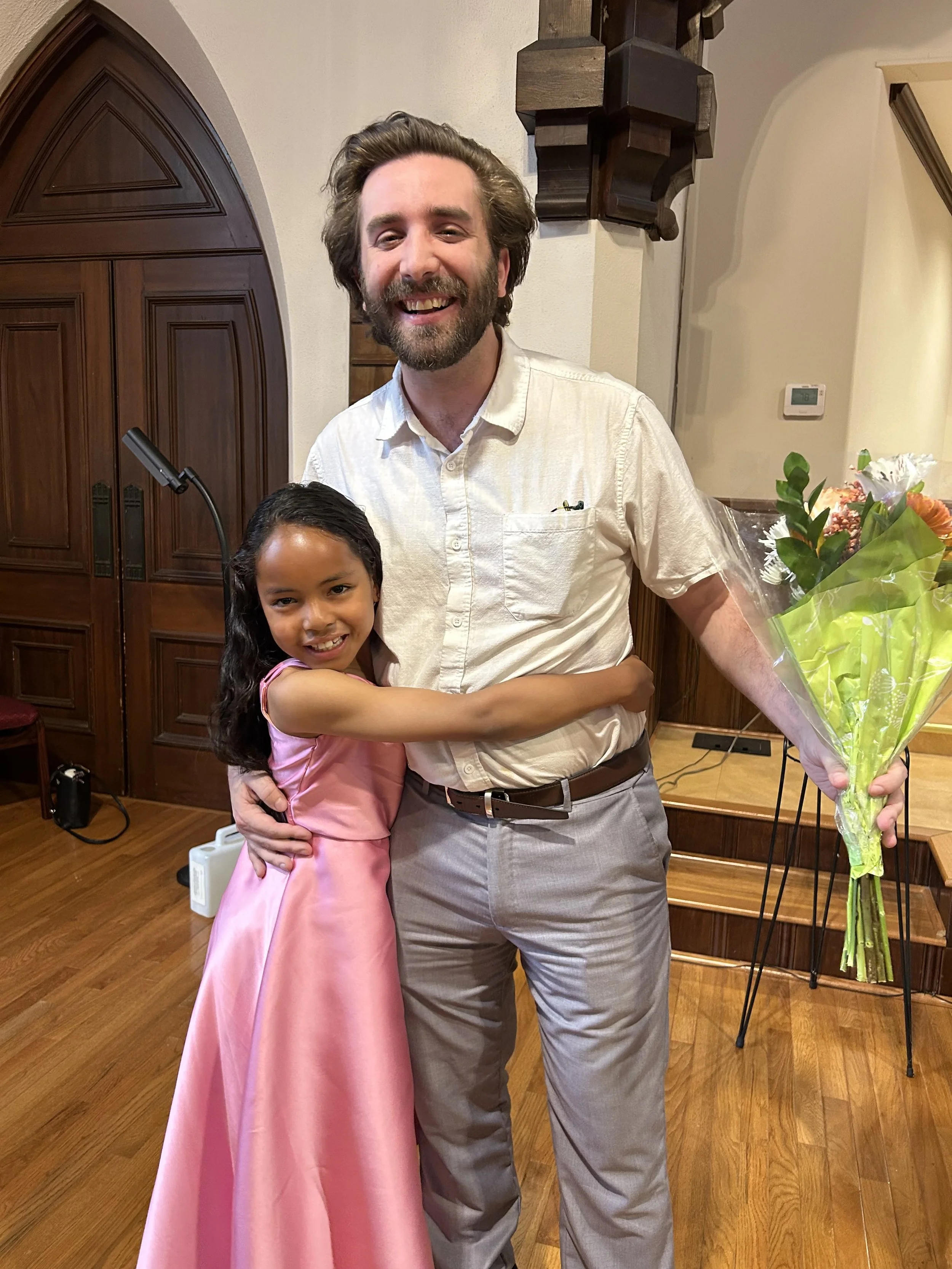 A man and a young girl are smiling and hugging in a room with wooden walls and floors. The man is holding a bouquet of flowers.