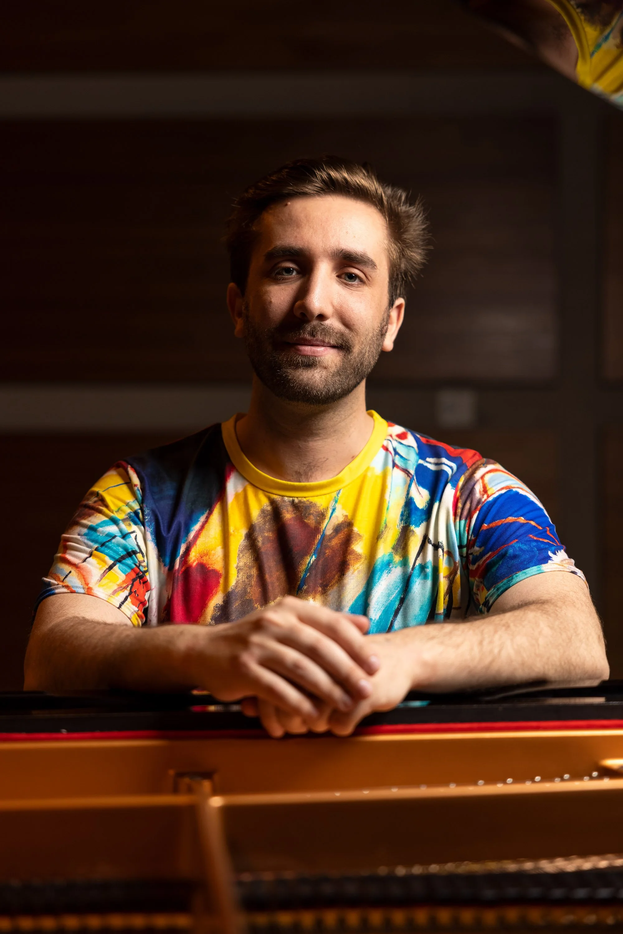 A man sitting at a piano, smiling, wearing a colorful, abstract patterned t-shirt, with dark wood paneling in the background.
