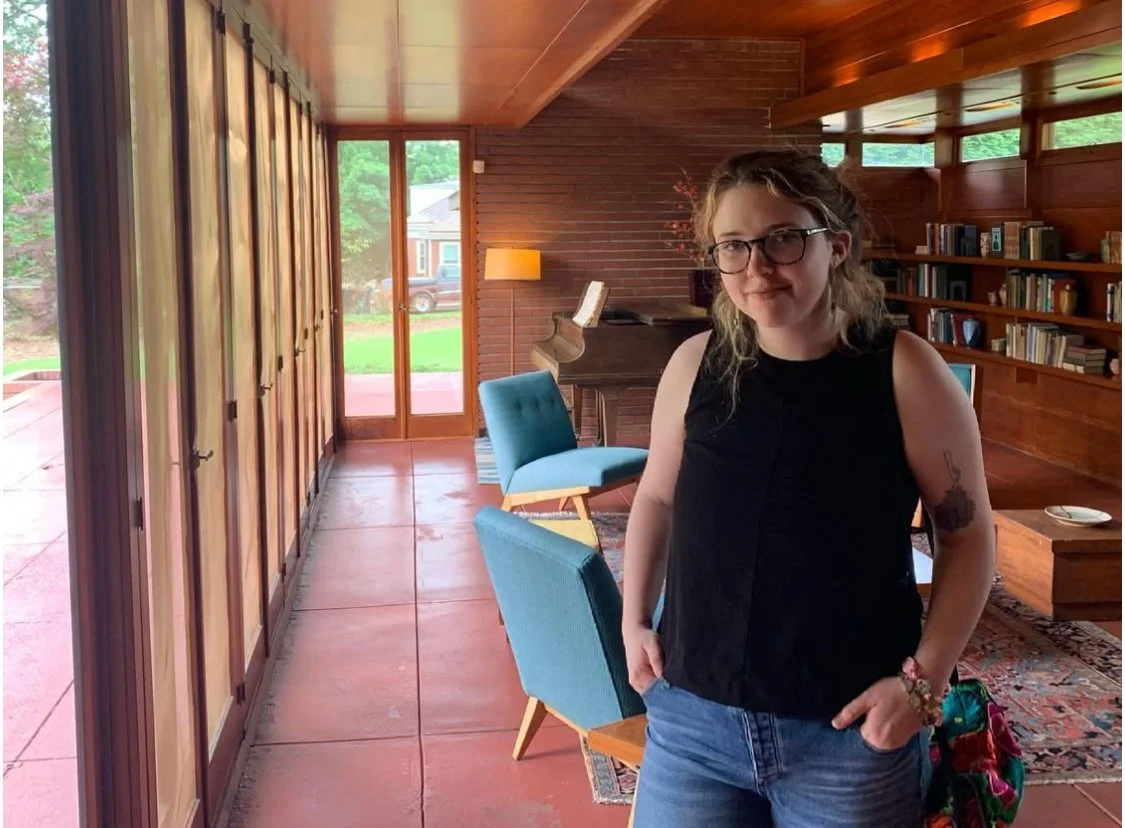 Callista, a white nonbinary person is standing to the right across a row of windows. Callista has wavy hair and glasses..