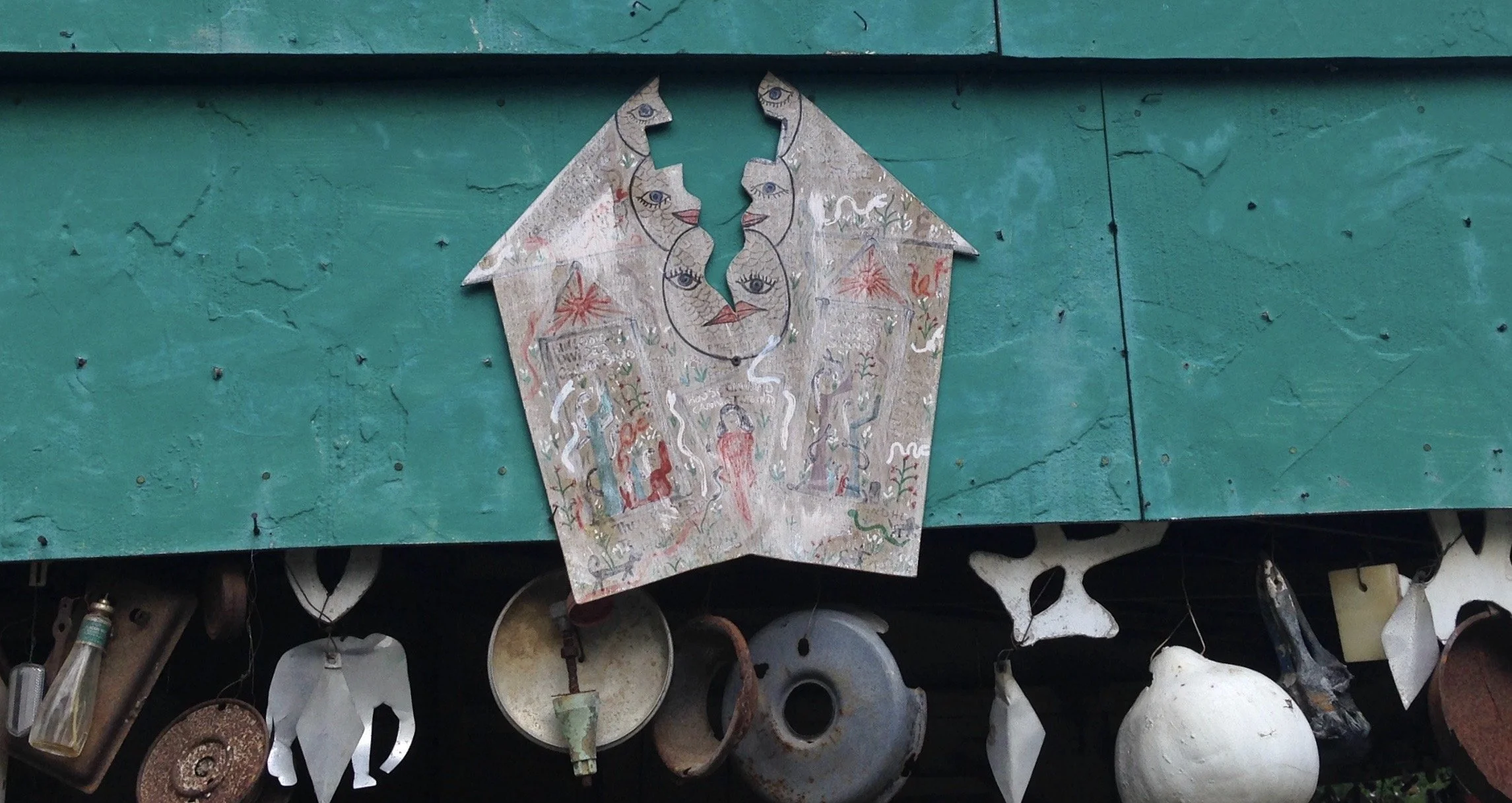 Artwork by Howard Finster of a house with faces on it being split down the middle. Behind is a teal piece of wood and rusty metal objects hanging underneath.