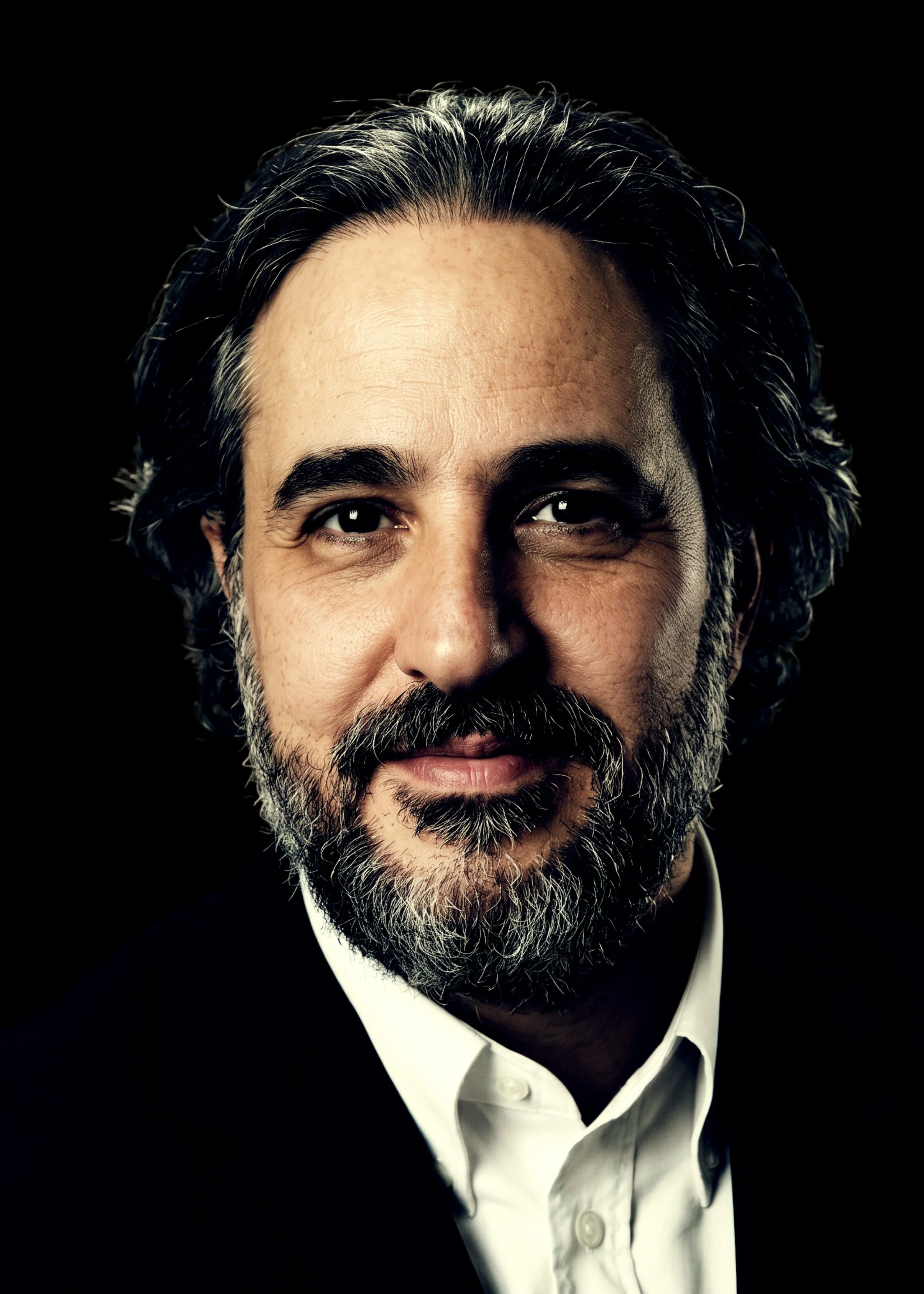 Close-up of a middle-aged man's face with gray hair and beard, wearing a black suit and white shirt, against a black background.