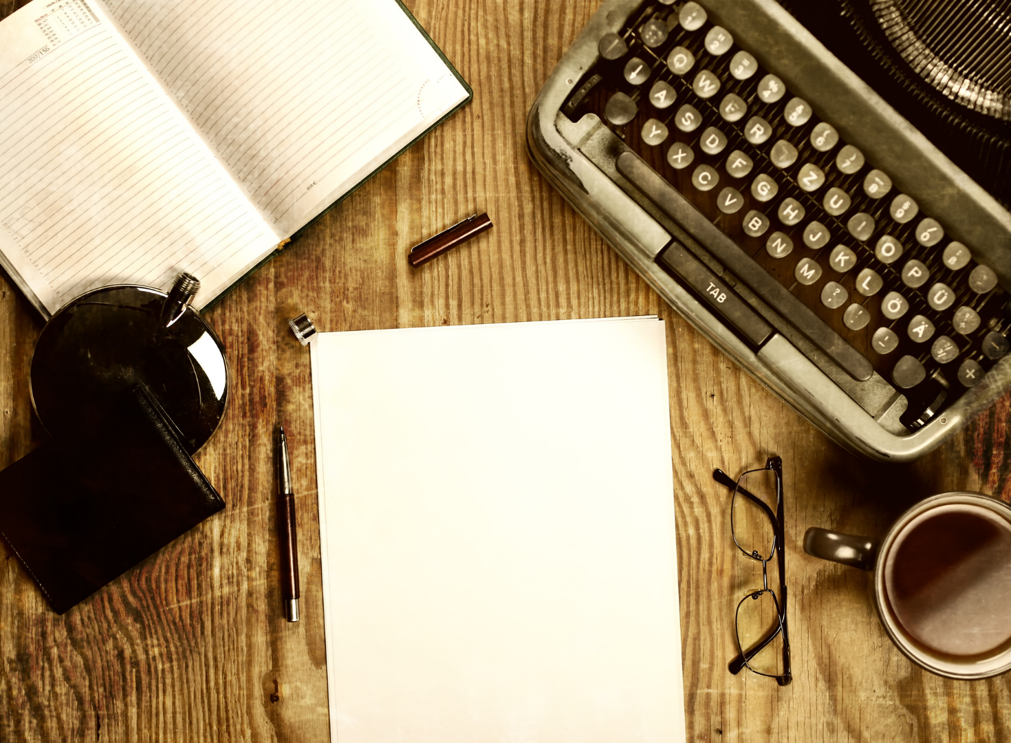 A wooden desk with an open notebook, a blank sheet of paper, a vintage typewriter, a pair of eyeglasses, a mug of coffee, a pen, a wallet, a small notebook, and a mirror.