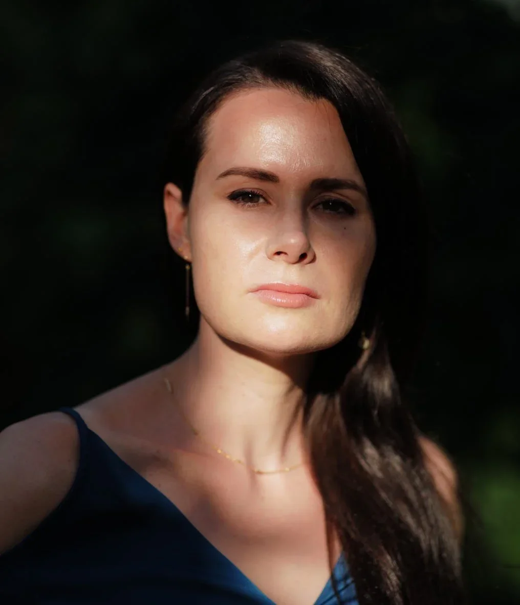 A woman with dark hair and light skin, wearing a dark blue top and gold jewelry, standing outdoors with sunlight on her face, against a dark background.