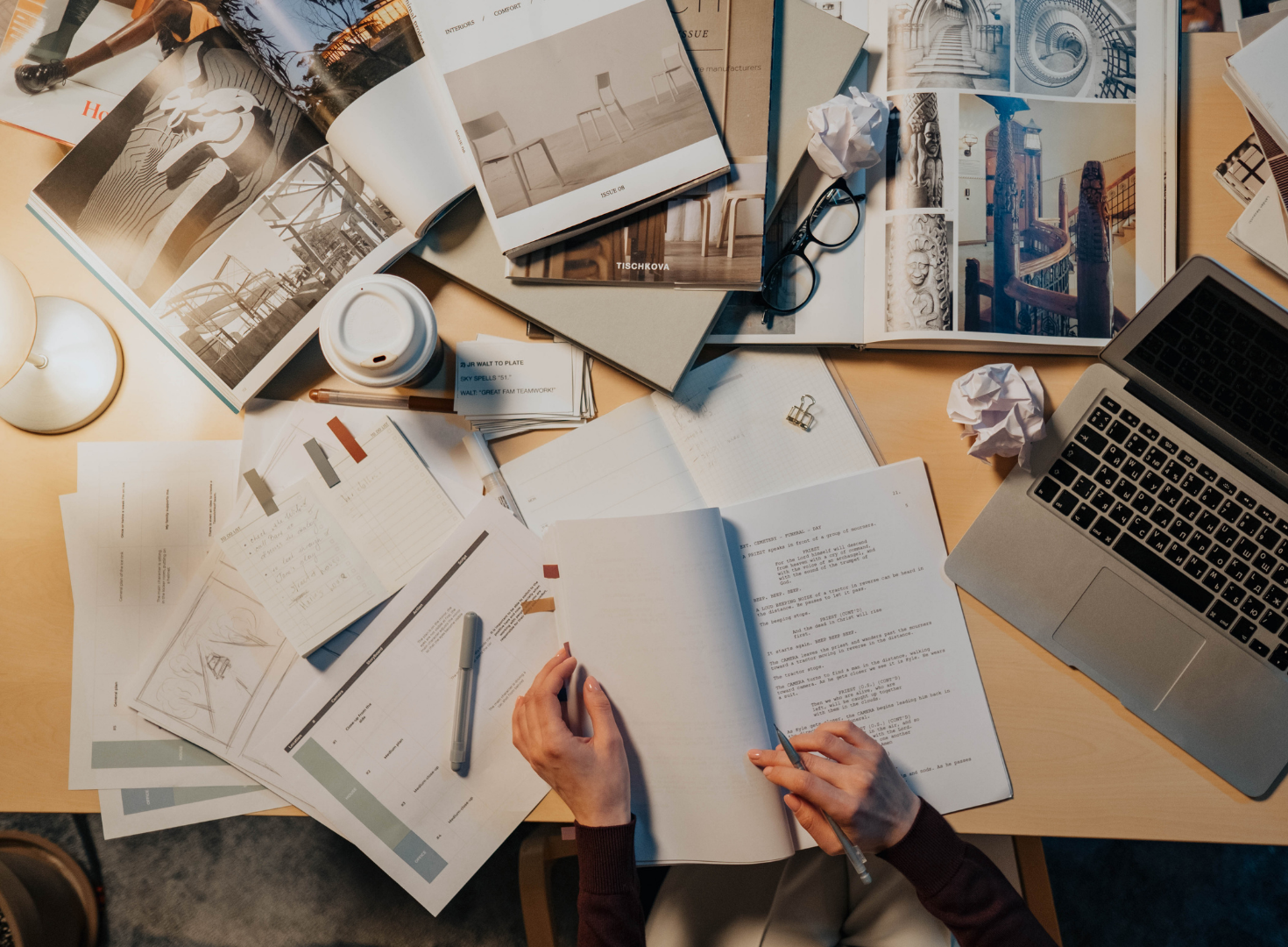 A cluttered desk with open magazines, notebooks, printed documents, a laptop, a coffee cup, a pen, crumpled tissues, glasses, and a binder clip, suggesting a workspace or study area.