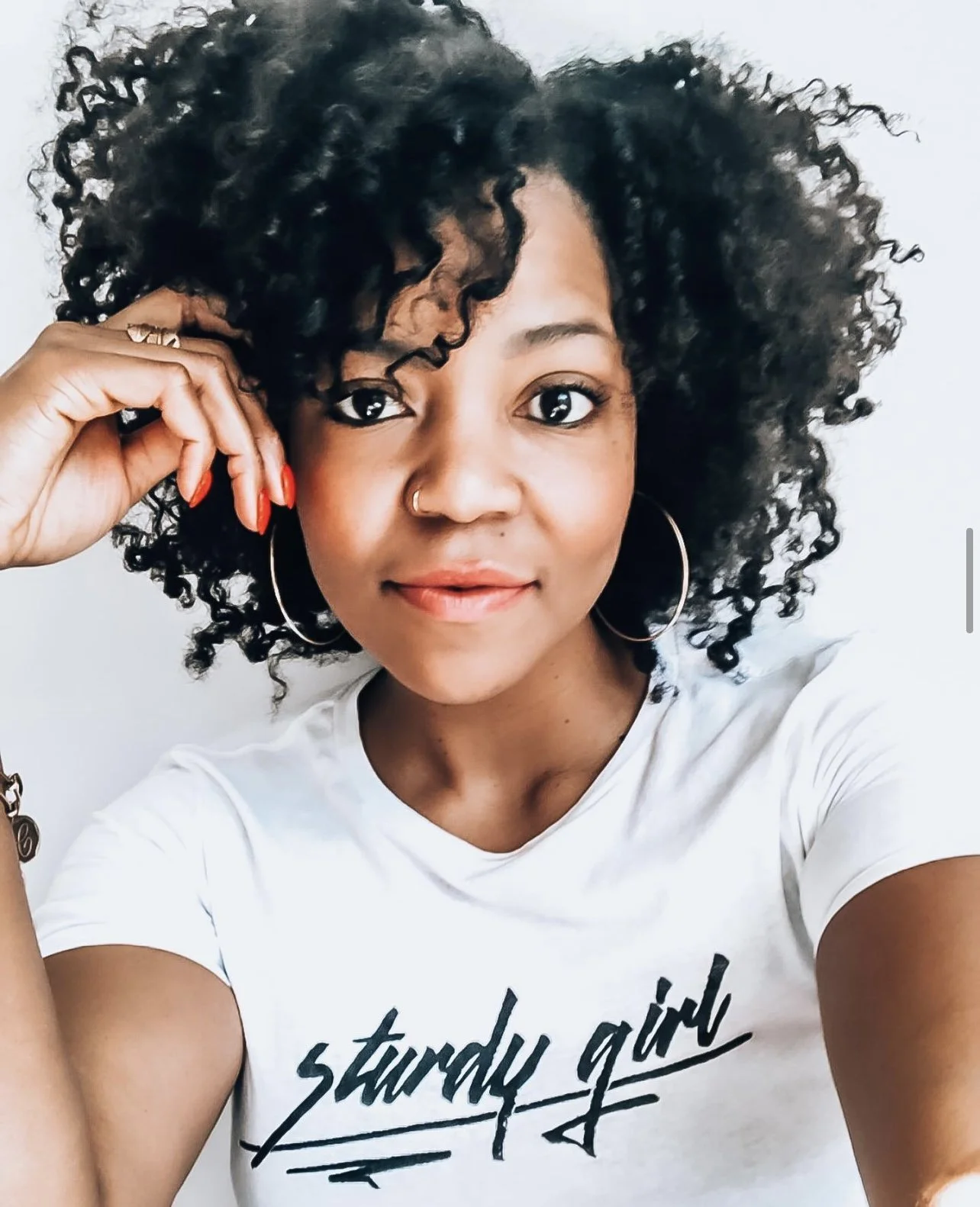 Chrissy King with curly black hair taking a selfie, wearing large hoop earrings, a nose ring, and a white t-shirt that says 'sturdy girl,' smiling with red lipstick and natural makeup.