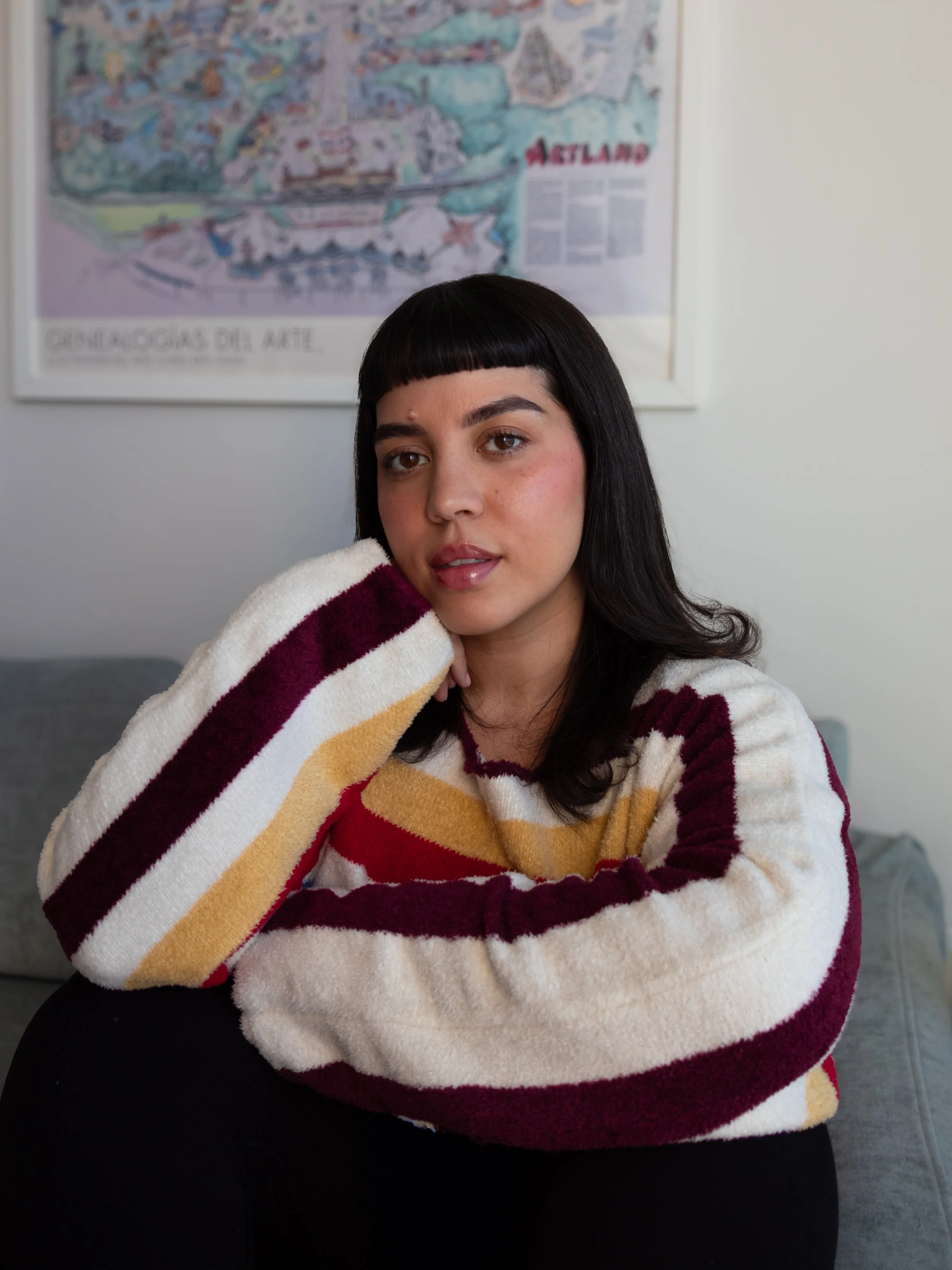 Melanie Santos with dark hair, wearing a colorful striped sweater, sitting on a gray couch, with a map of Alesund in the background on a white wall.