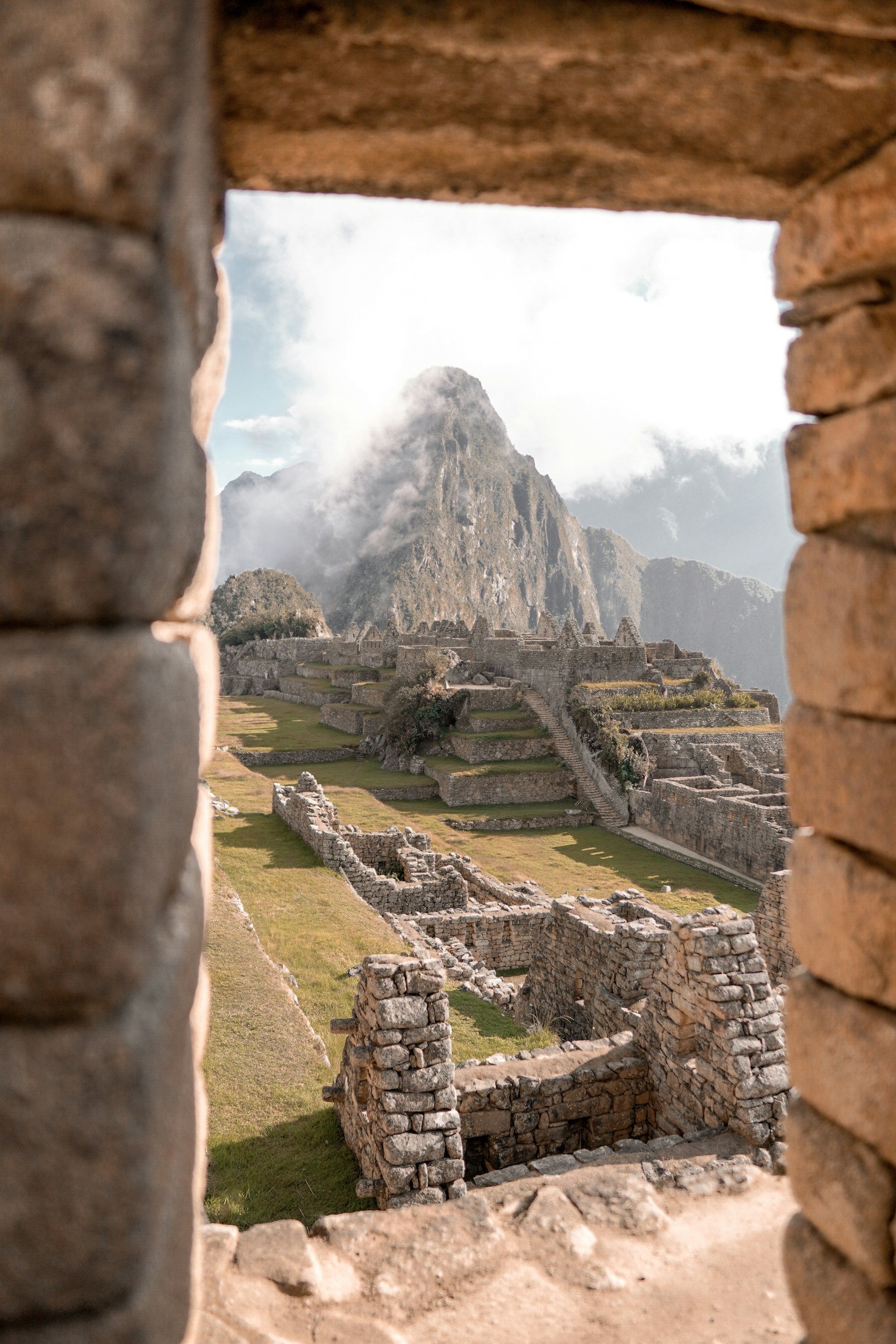 View of Machu Picchu with ancient stone ruins and terraces, seen through a rectangular window in a brick wall, with mountain peak and cloudy sky in the background.