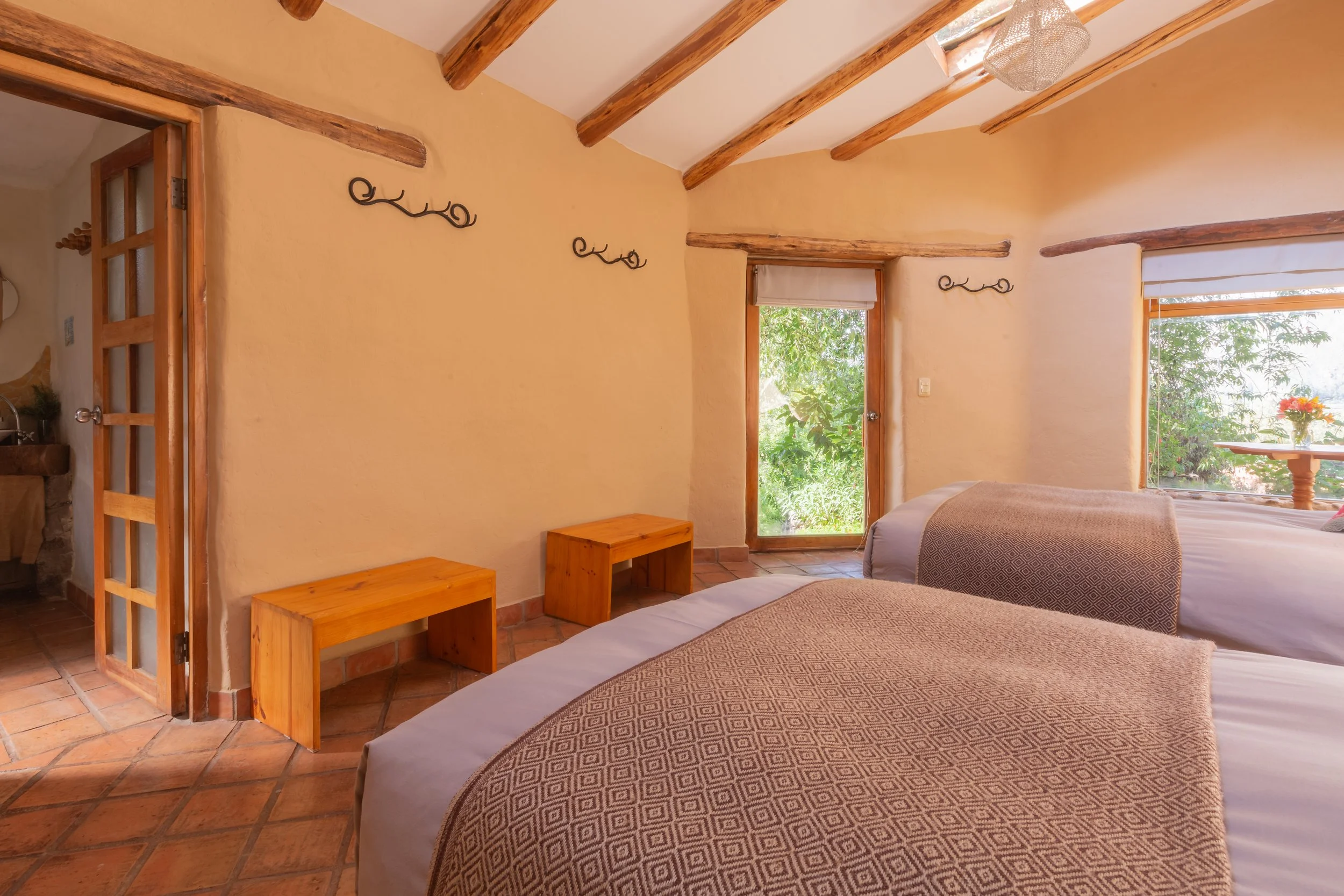 A cozy bedroom with two beds, each with geometric patterned bedspreads. The room has beige walls, wooden ceiling beams, and natural light from two windows with white blinds, one showing greenery outside. There's a small wooden nightstand and decorati