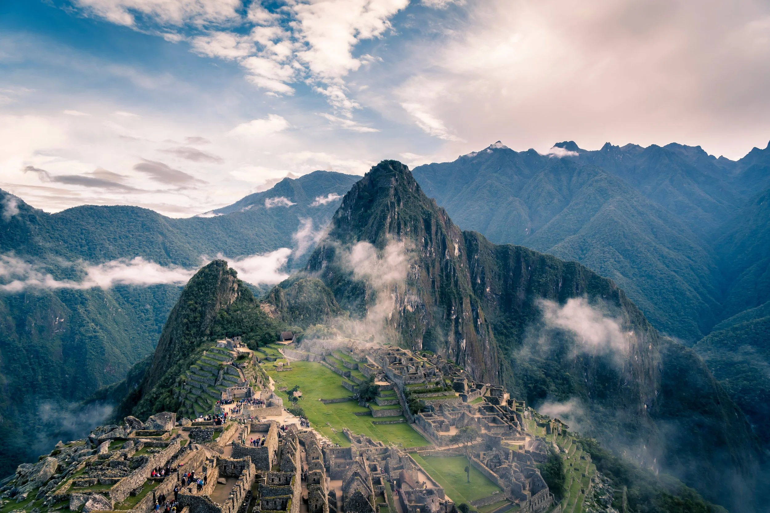 Aerial view of Machu Picchu, the ancient Incan city, surrounded by lush green mountains and clouds, with tourists exploring the archaeological site.