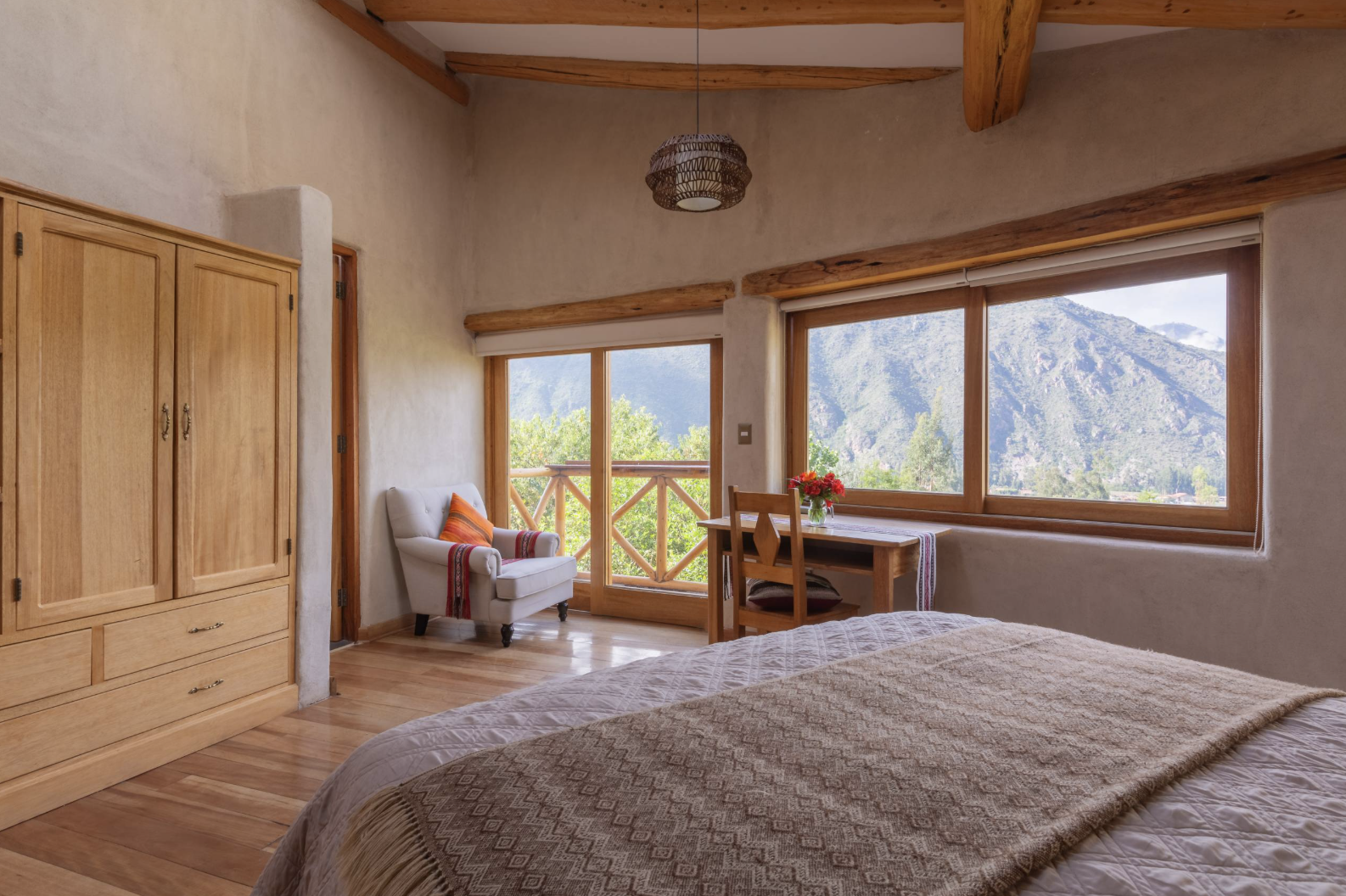 A cozy bedroom with a large window showing a mountainous landscape, a bedside table with a vase of red flowers, a small sitting area with a white armchair and colorful pillow, a wooden wardrobe, and warm wooden furniture.