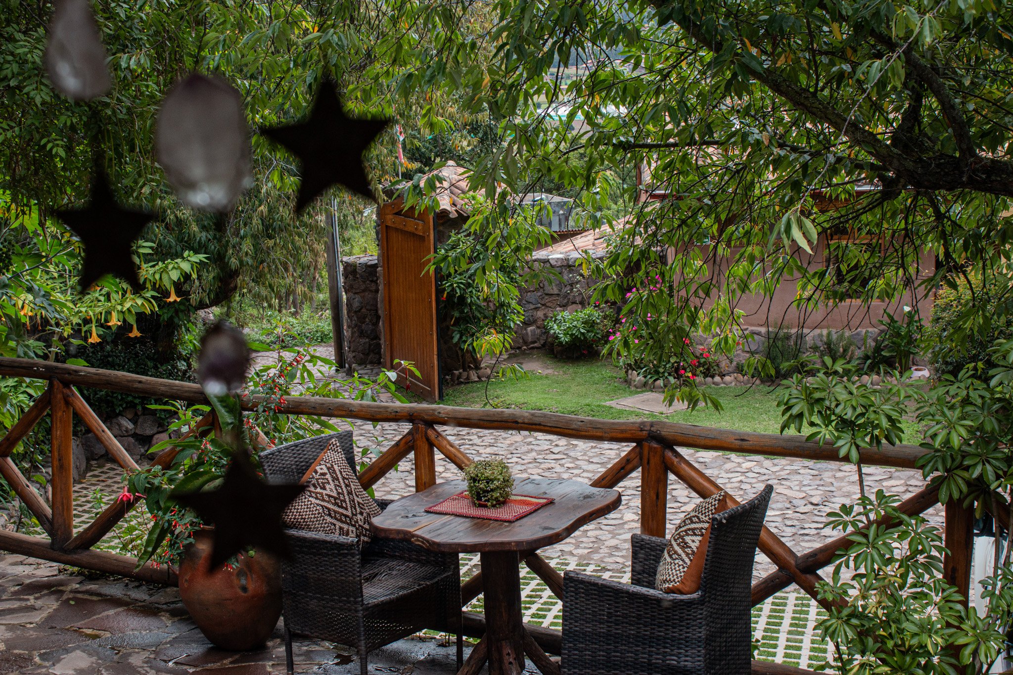 A cozy outdoor patio with a wooden table and four wicker chairs with patterned cushions, surrounded by lush green trees and flowering plants, with a stone pathway and a stone wall in the background.