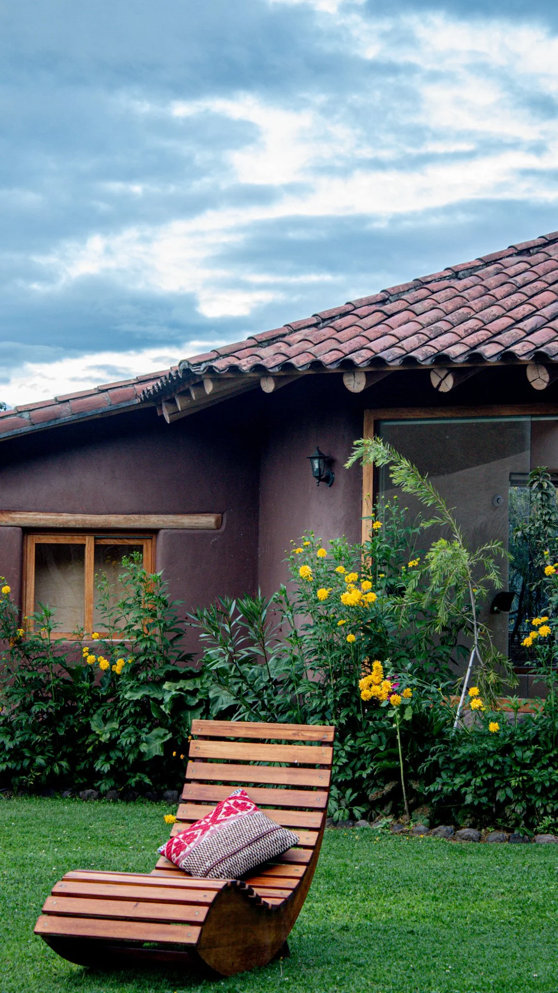 A backyard scene with a wooden lounge chair on a grassy lawn, decorated with cushions, in front of flowering plants and a house with a sloped terracotta tile roof and window. The sky is partly cloudy.