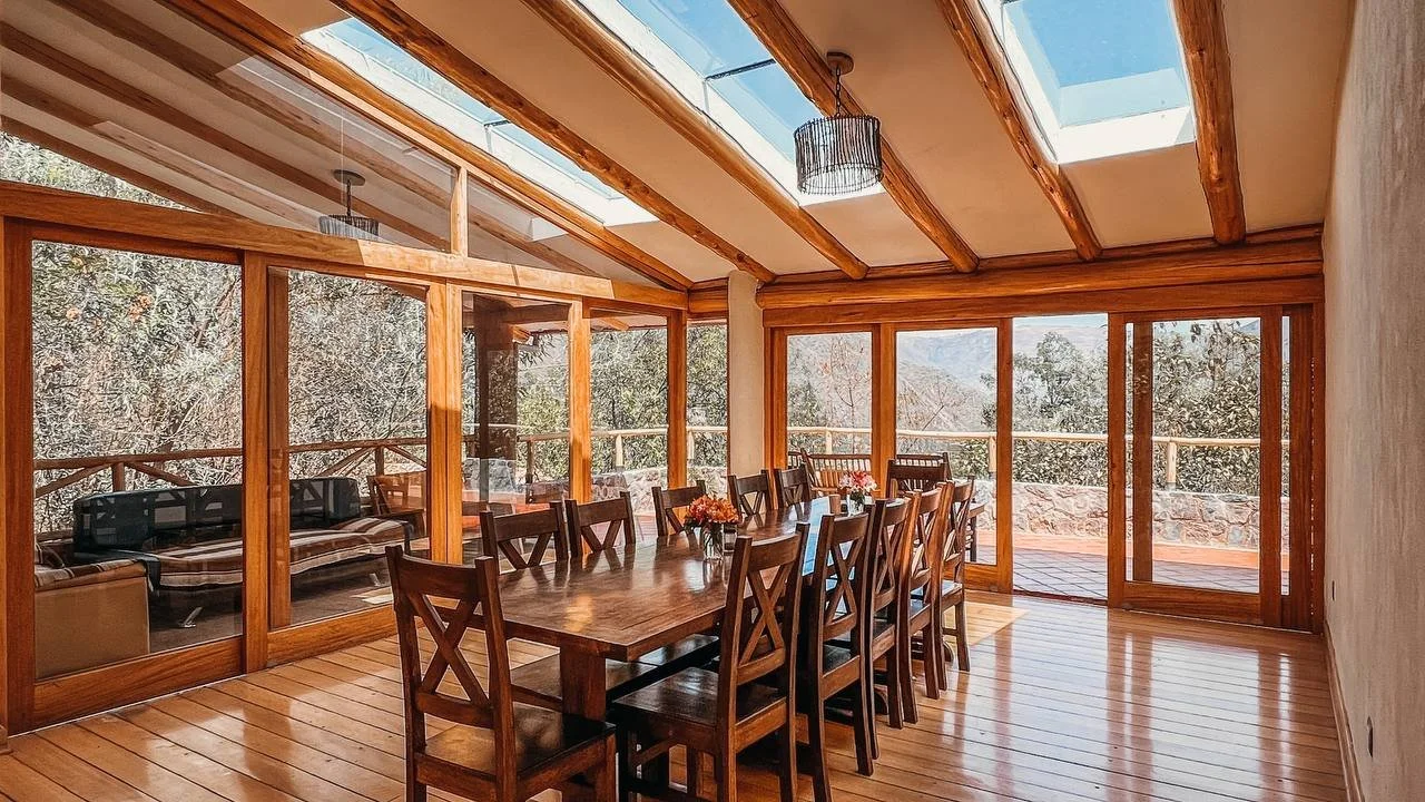 A sunlit dining room with a large wooden table surrounded by matching chairs, and large glass sliding doors and windows providing a view of trees outside. The room has a wooden floor and ceiling beams.