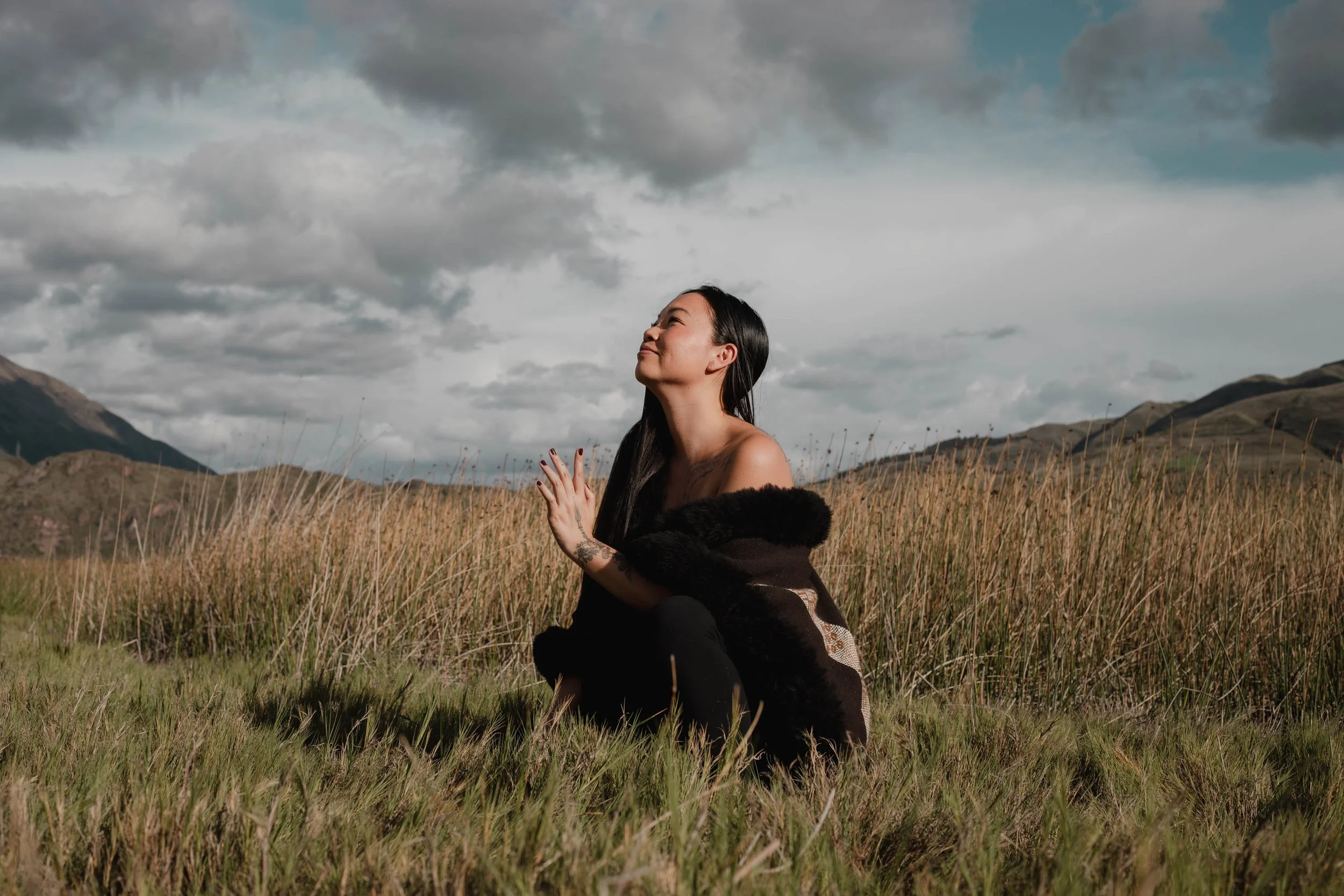 Tara Tao Center of Destiny with long dark hair and tattoos on her arm kneels in a field of tall grass, smiling gently with her eyes closed, against a backdrop of mountains and a cloudy sky.